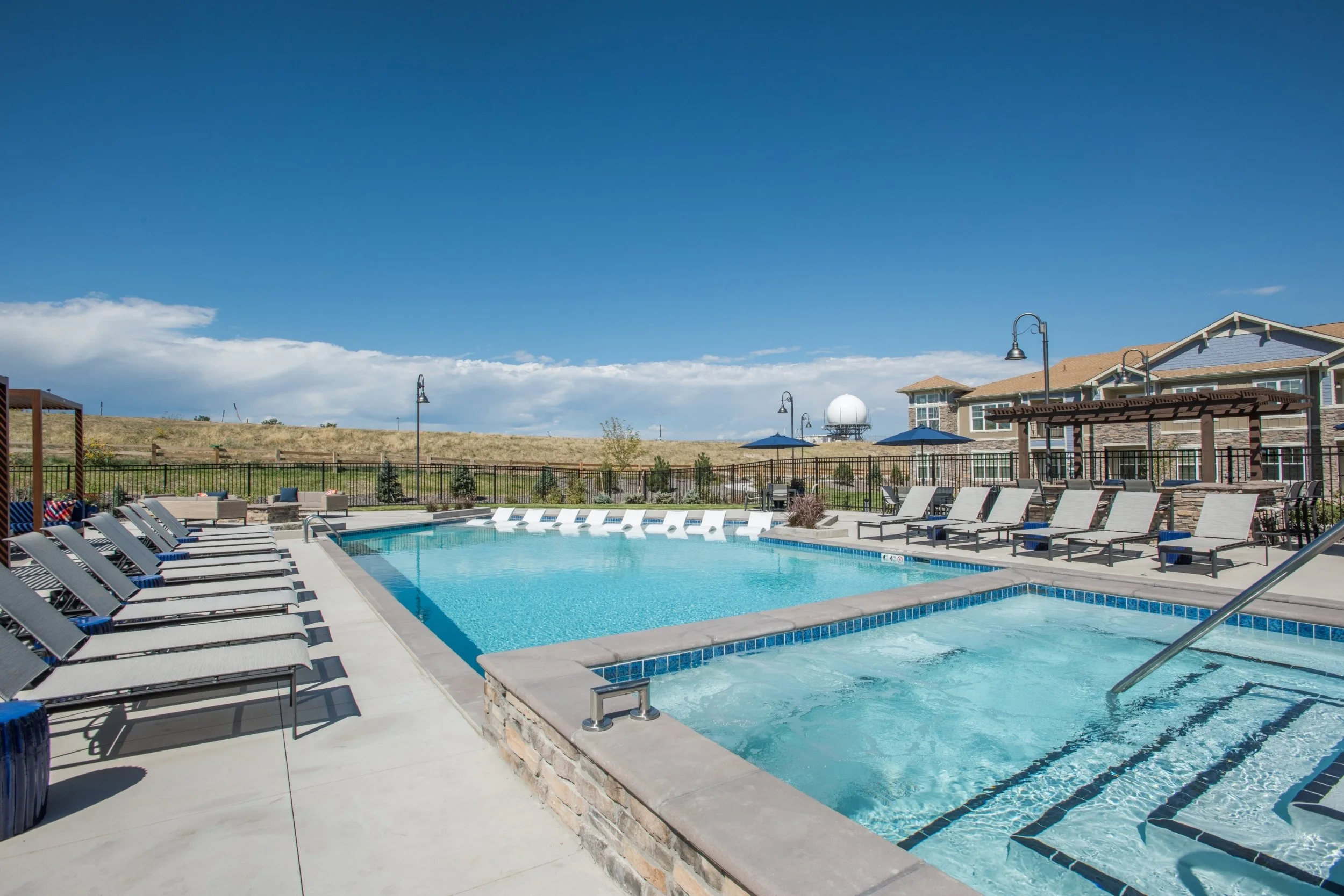 An outdoor swimming pool area with lounge chairs, umbrellas, and a hot tub under a clear blue sky.