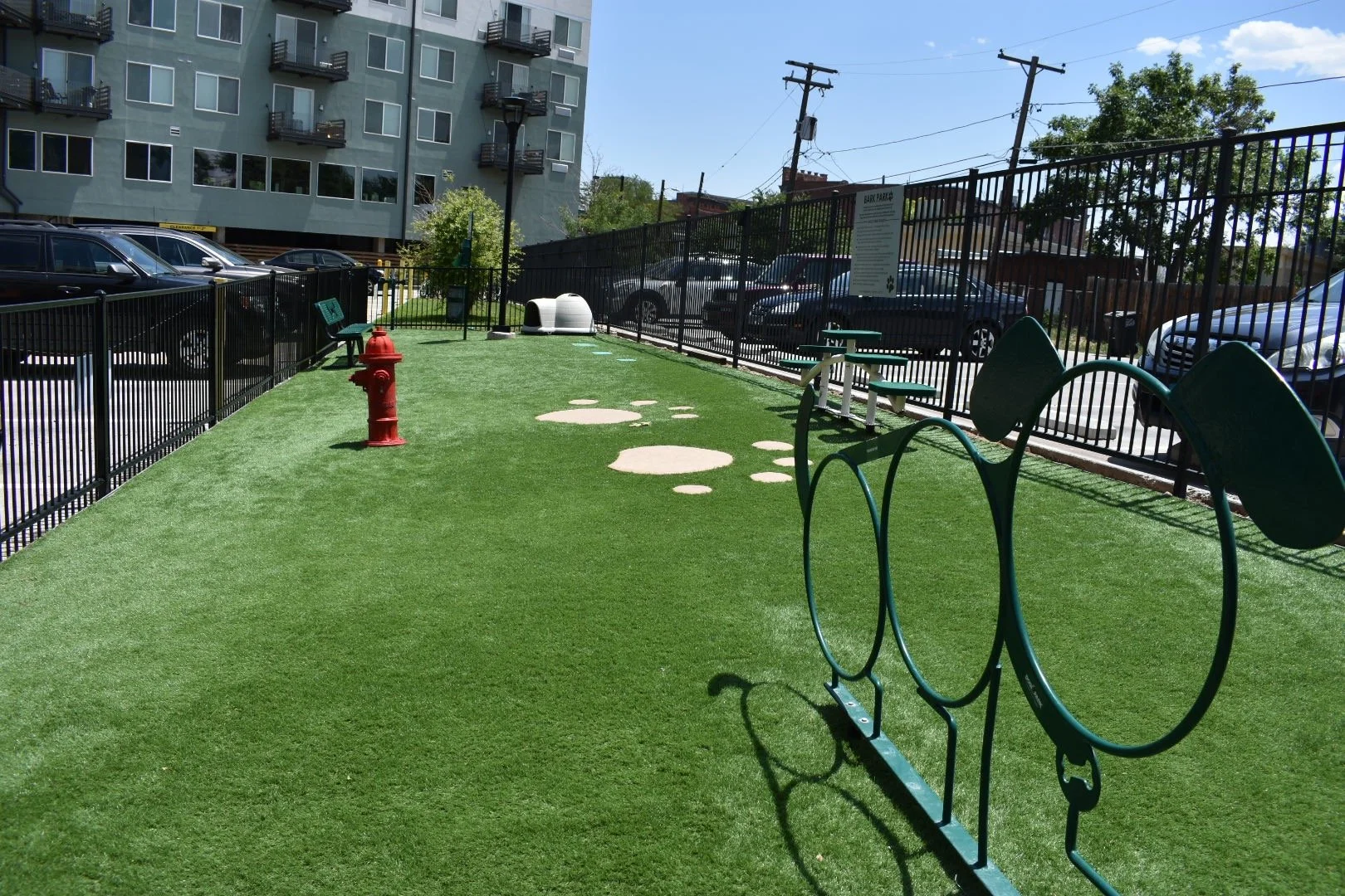Apartment complex outdoor dog park with artificial grass, street lamp, benches, and parking lot in background.