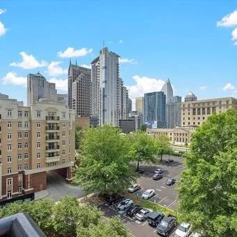 View of a city skyline with modern skyscrapers, a parking lot, and green trees under a partly cloudy sky.