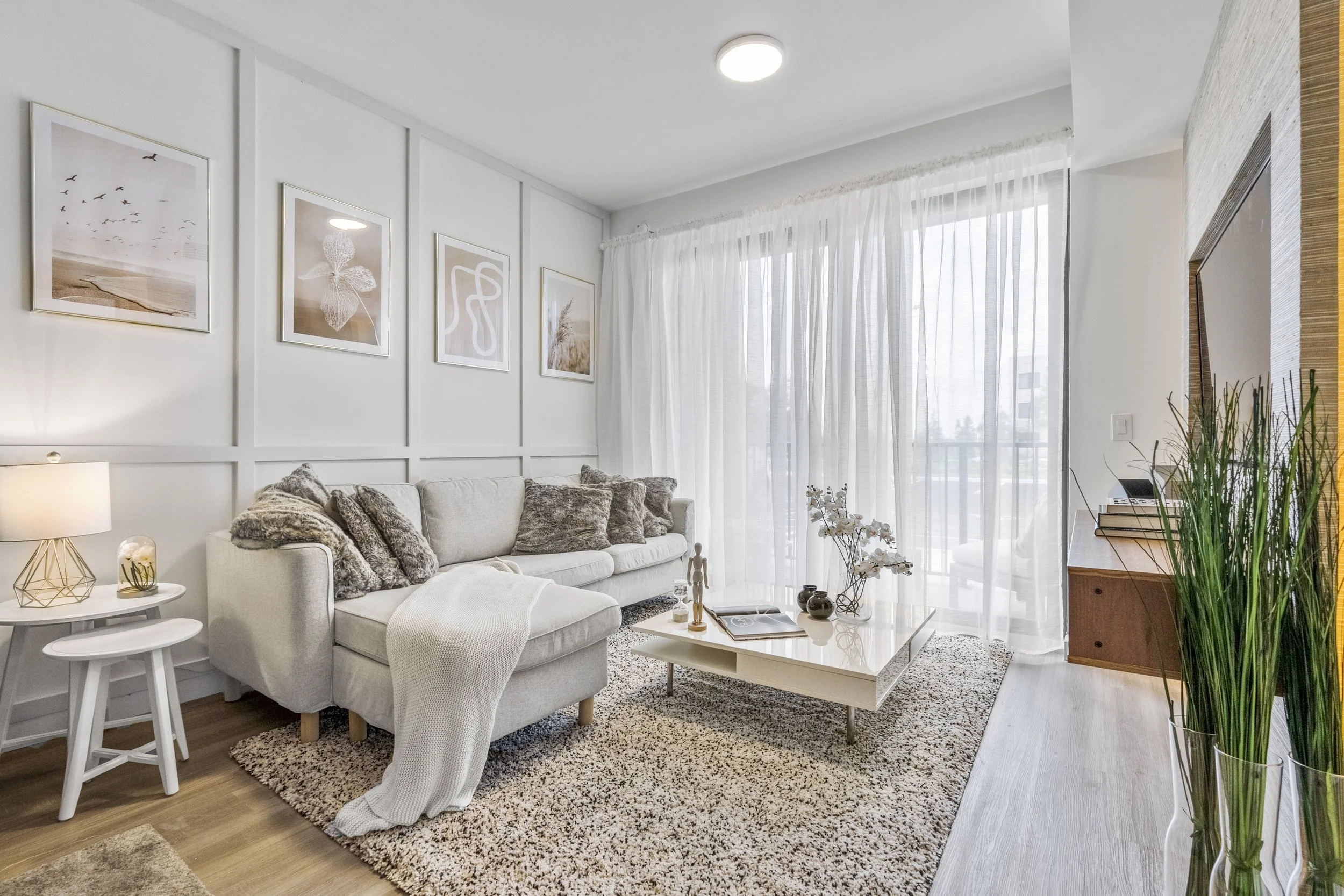 Bright living room with white sectional sofa and plush pillows, white coffee table with decorative items, art prints on the wall, sheer white curtains, wooden TV stand, and large green plant.