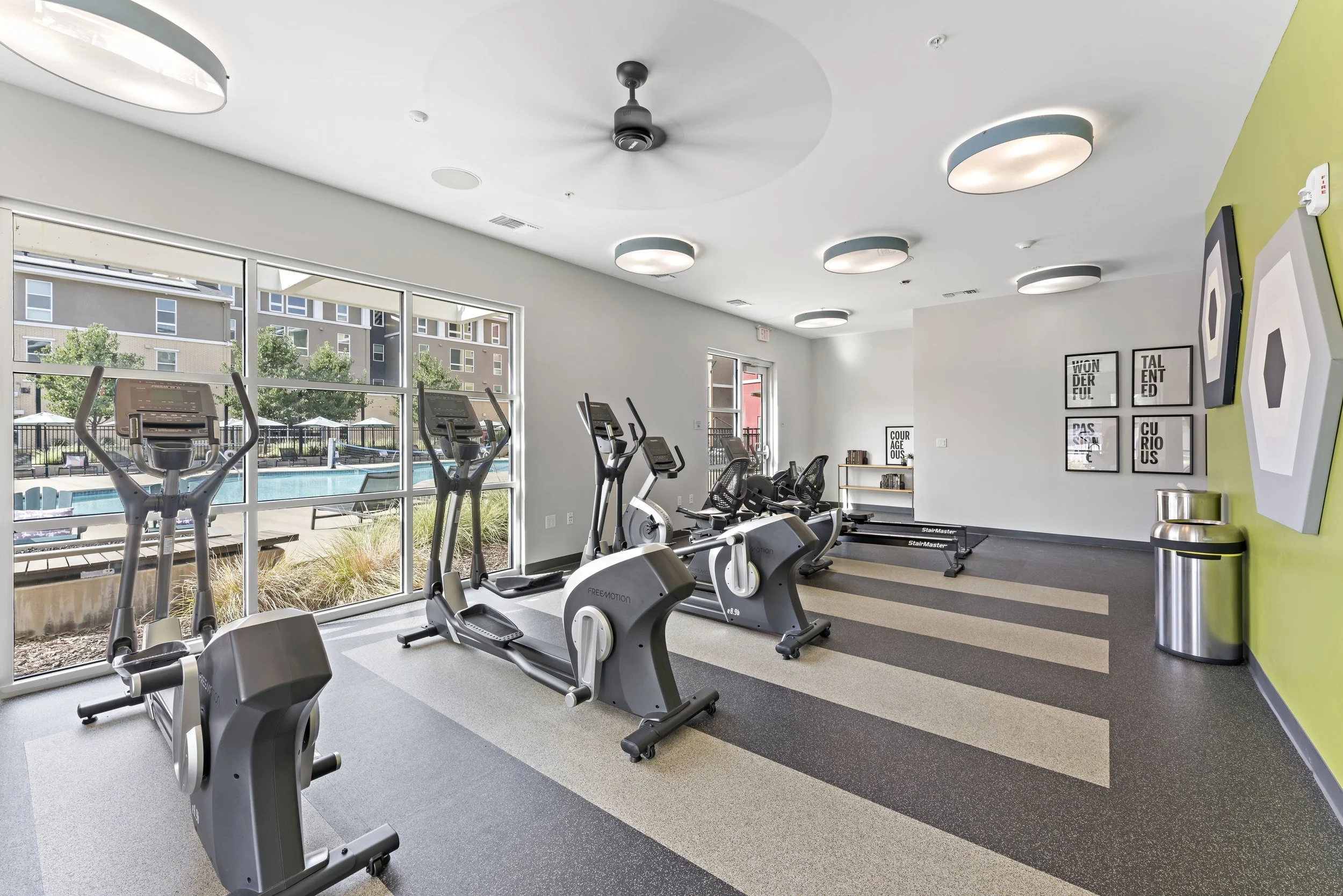 Indoor gym with several cardio machines facing large windows overlooking swimming pool and surrounding apartments, decorated with wall art and modern lighting.