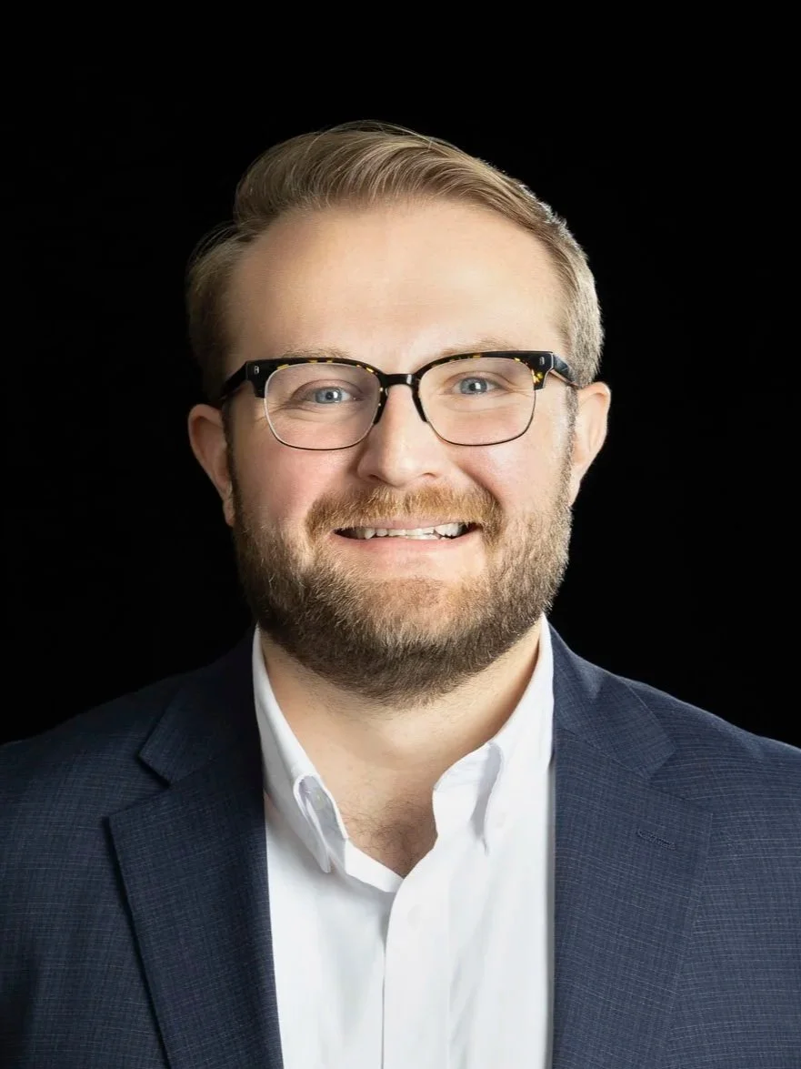 Professional portrait of a man with blonde hair, glasses, and a beard, smiling, wearing a navy blazer and white shirt against a black background.