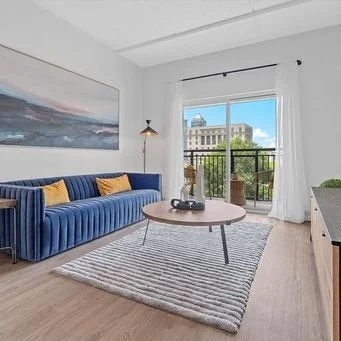 Living room with blue velvet sofa, yellow pillows, framed landscape art, round coffee table, striped rug, floor lamp, and sliding glass door leading to a balcony