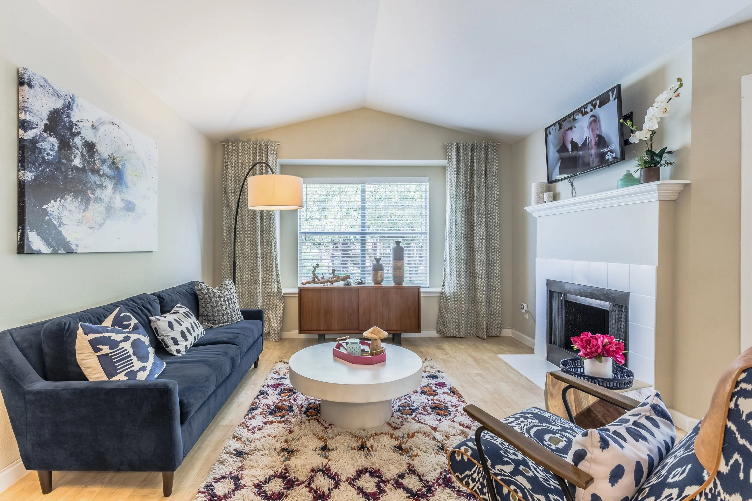 Living room with a navy sofa, patterned cushions, a white round coffee table, a colorful rug, a wooden sideboard, a window with curtains, a wall-mounted TV, a white fireplace with pink flowers, and a blue and white armchair.