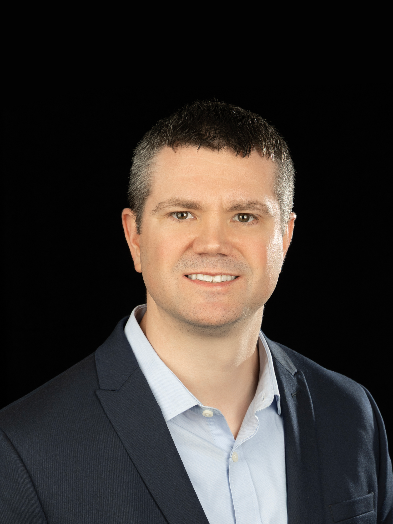 Headshot of a man with short dark hair, wearing a dark blazer and light blue collared shirt, smiling against a black background.