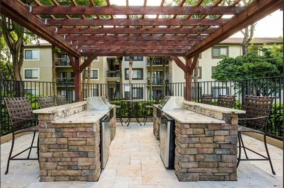 Outdoor rooftop patio with two brick bar counters, bar stools, and a wooden pergola, surrounded by apartment buildings and greenery.