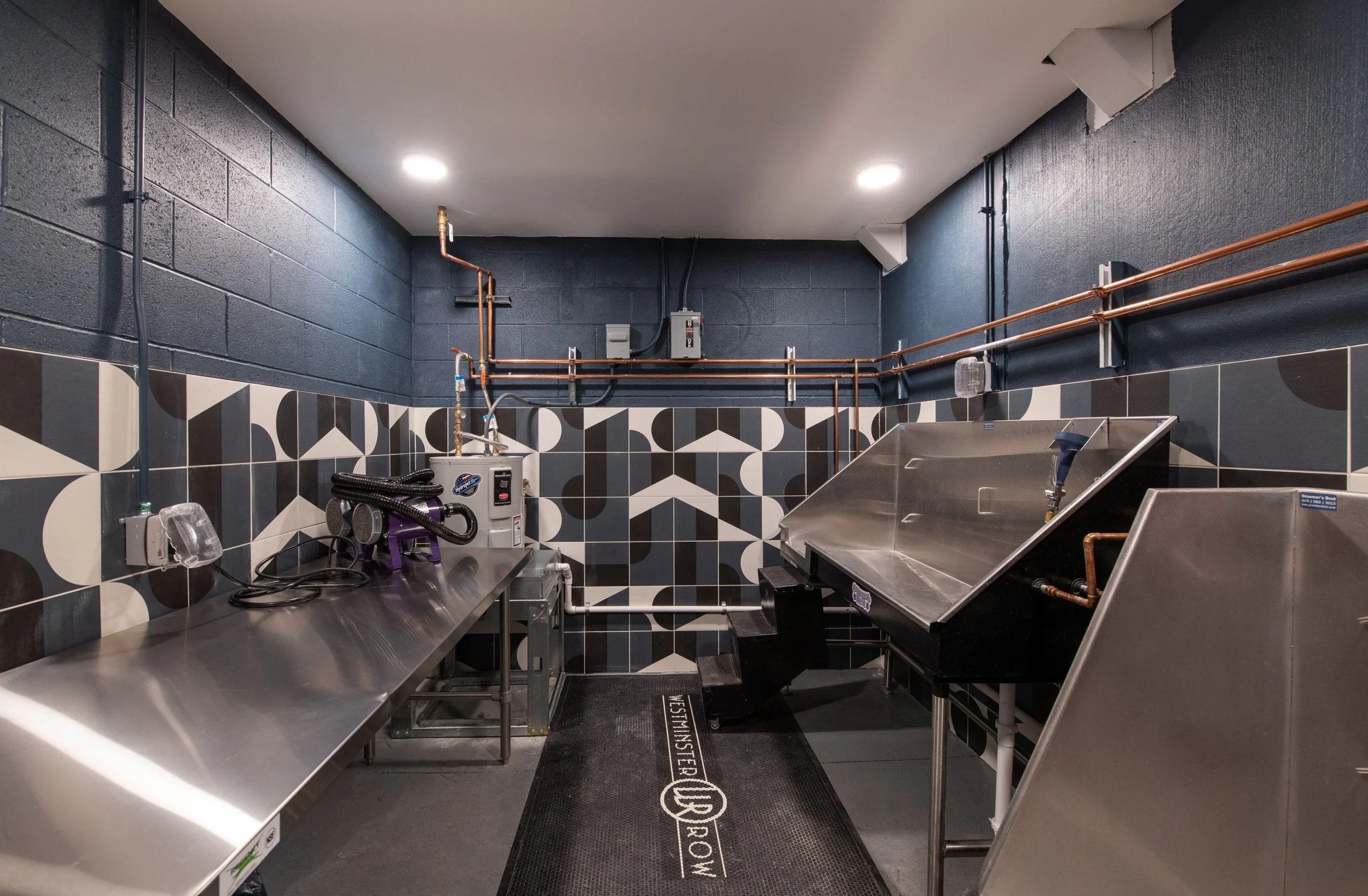 A stainless steel pet grooming station with a sloped sink on the right, a counter with grooming equipment on the left, and black and white patterned tiles on the wall. Copper pipes are mounted on the dark blue wall above.