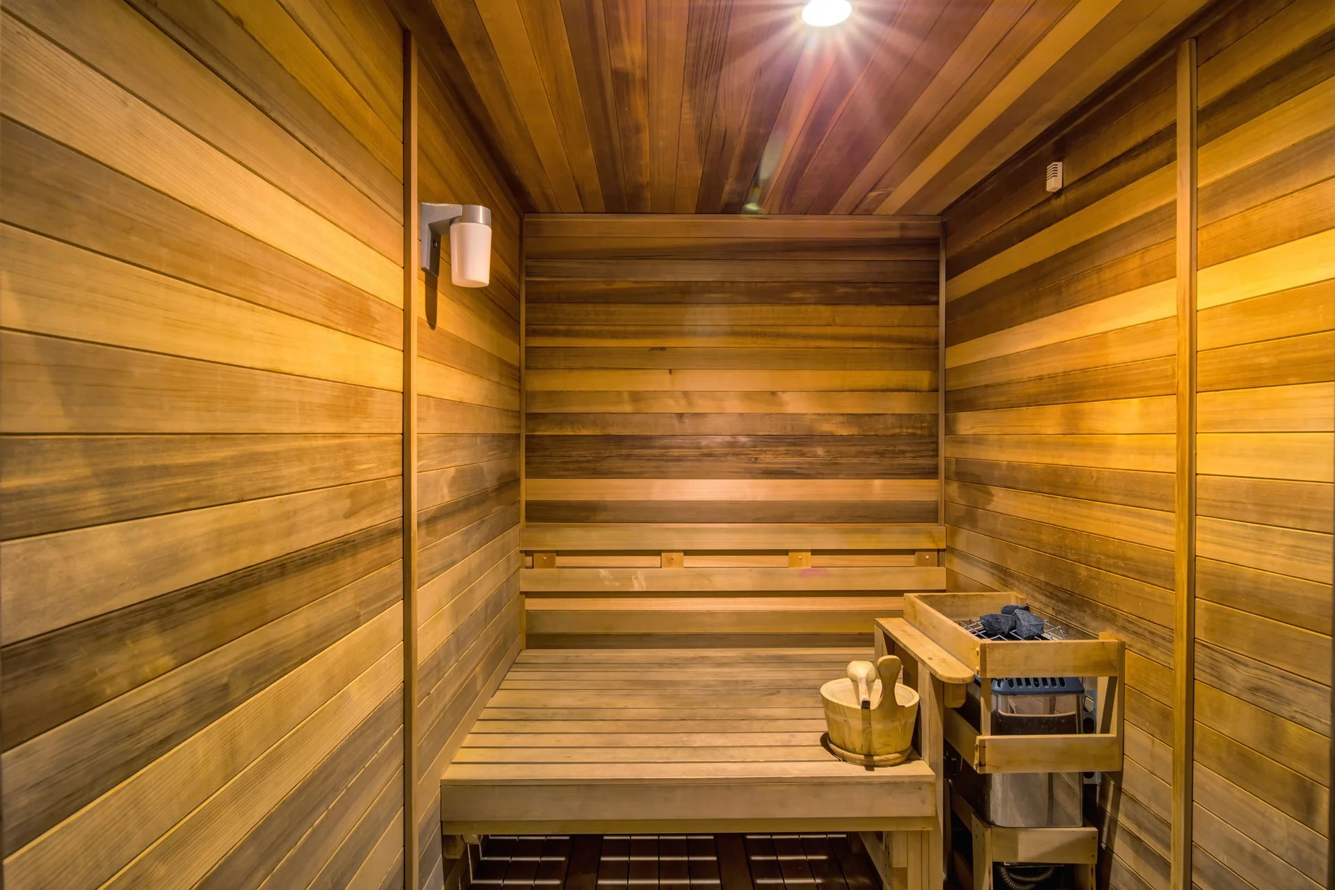 Inside a traditional wooden sauna with wooden benches, a heater with stones, a wooden bucket with ladles, and a wall-mounted light fixture.