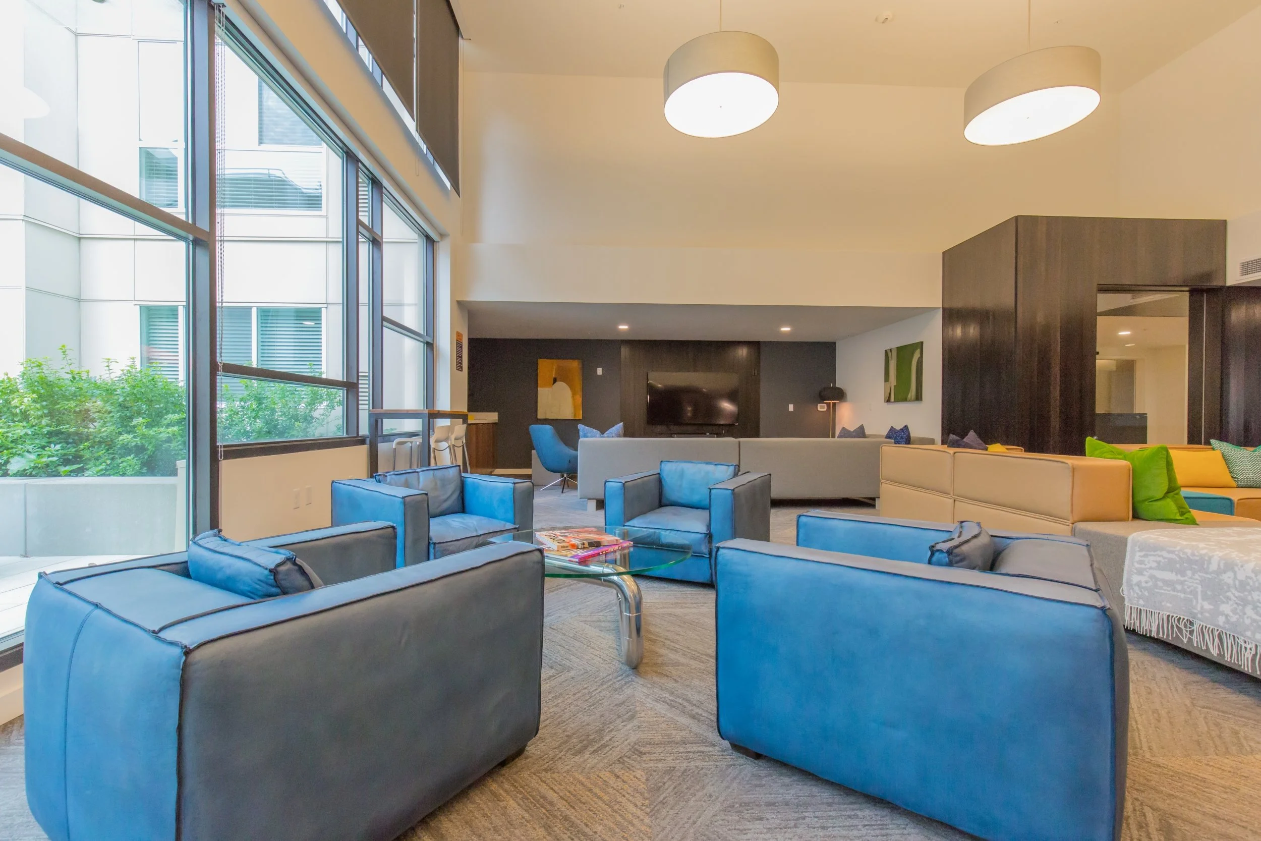 A spacious modern lounge area with several blue and beige sofas, a glass coffee table, large windows with greenery outside, a wall-mounted TV, and contemporary lighting fixtures.