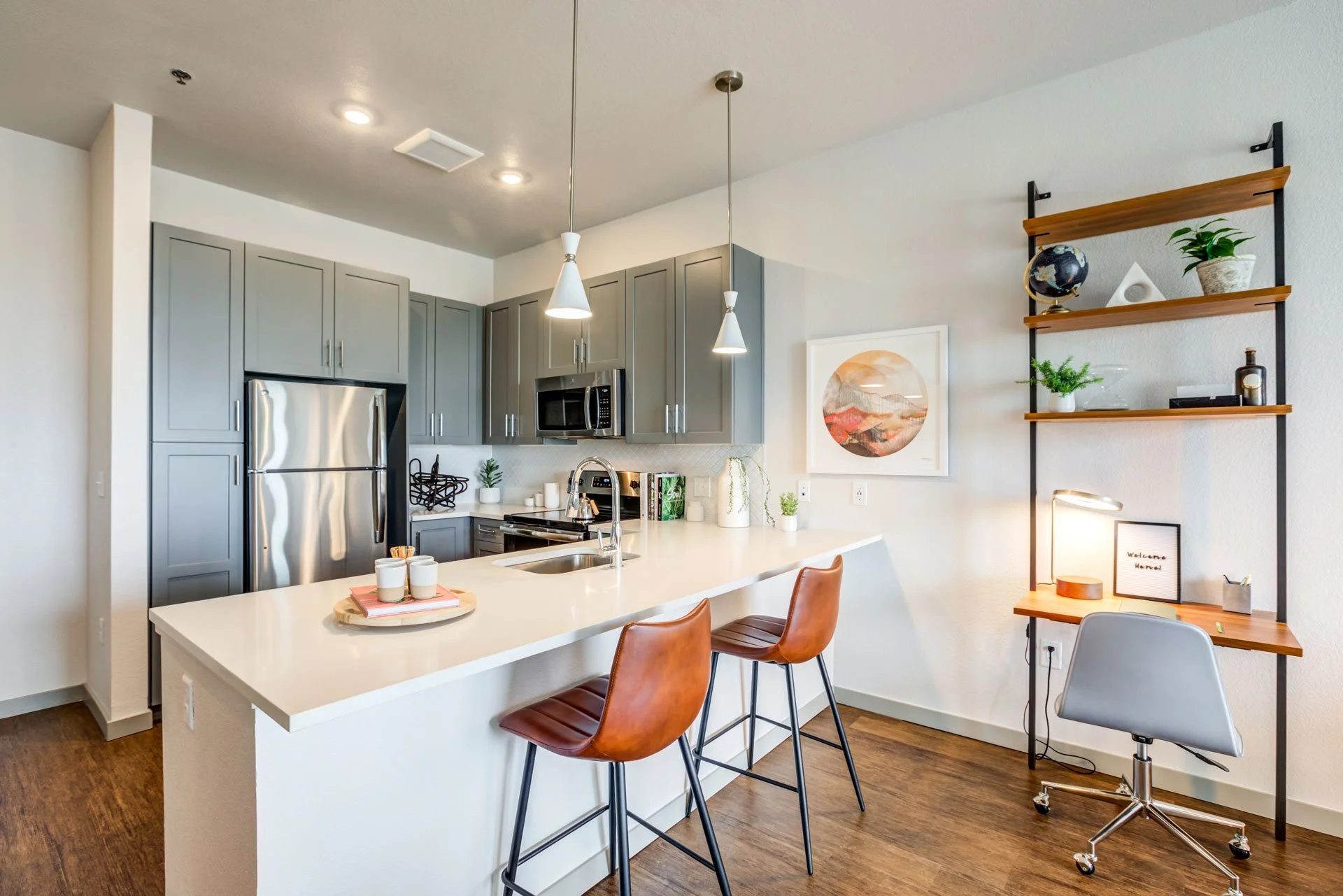 Modern kitchen with gray cabinets, stainless steel appliances, a white island with two brown chairs, a small desk with a gray chair, and decorative plants and artwork.