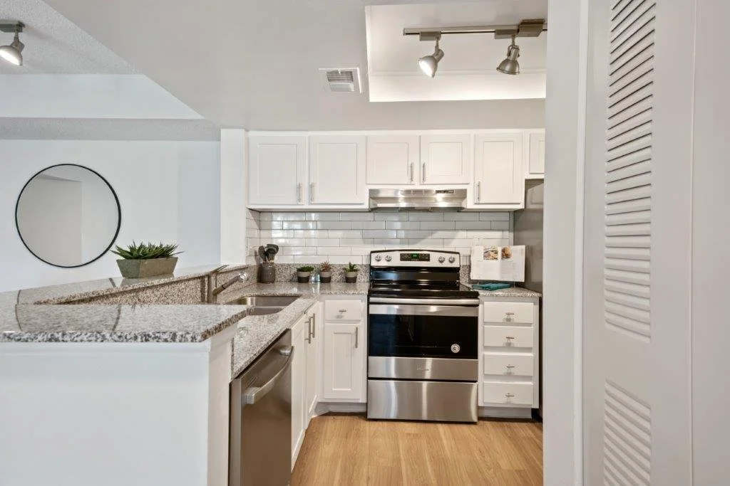 Kitchen with white cabinets, stainless steel stove, granite countertops, white subway tile backsplash, small potted plants, and a window or mirror with a round black frame.