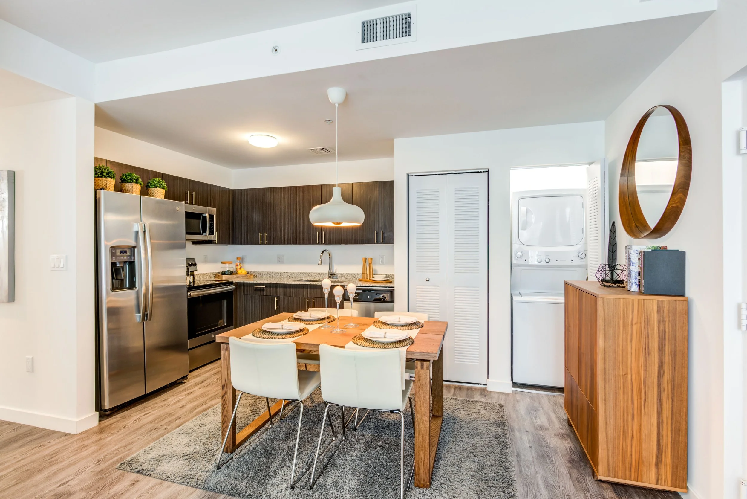 Modern kitchen and dining area with wood cabinetry, stainless steel refrigerator, white dining table, white chairs, and decorative items.