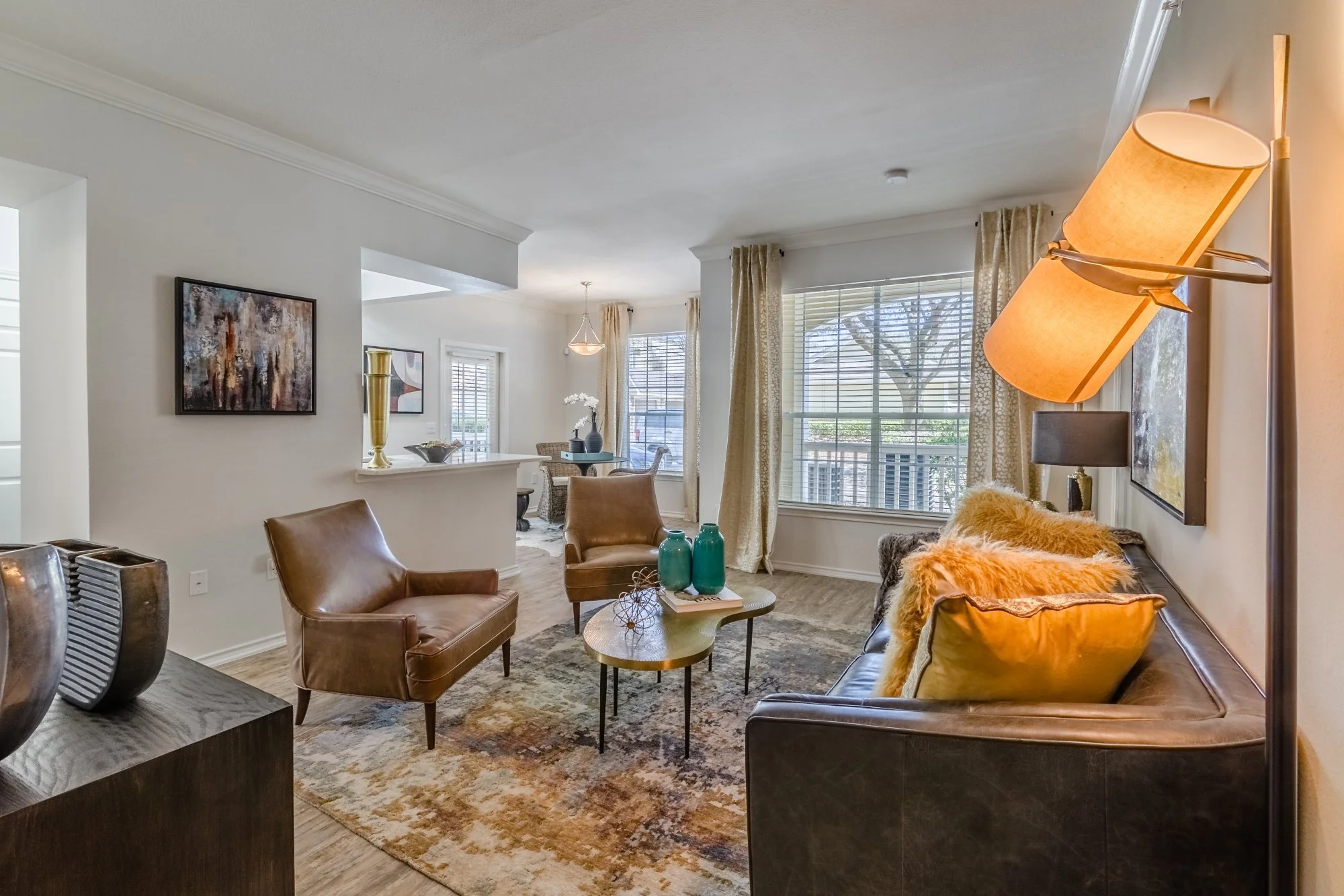 Living room with beige curtains, a leather sofa with furry pillows, a wooden coffee table with teal vases, leather armchairs, and large windows with blinds.