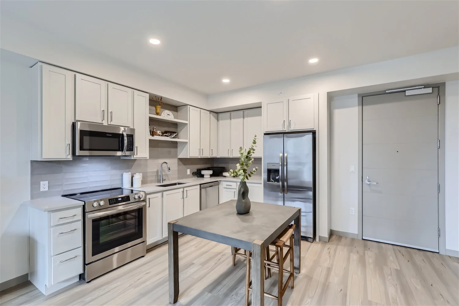Modern kitchen with white cabinets, stainless steel appliances, a wooden dining table with a vase, and light wood flooring.