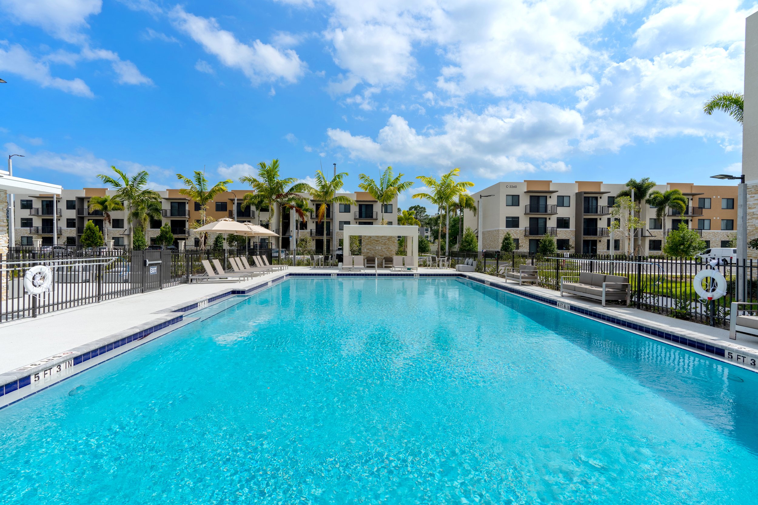 A swimming pool in a residential complex with blue water, surrounded by lounge chairs and palm trees under a partly cloudy sky.