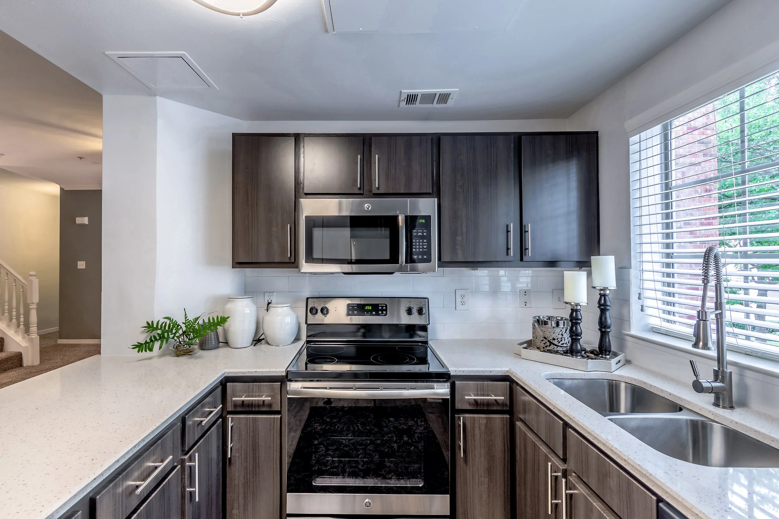 Modern kitchen with dark wood cabinets, white speckled countertops, stainless steel microwave and oven, double sink near window with blinds, decorative candle holders, and white vases on the counter.