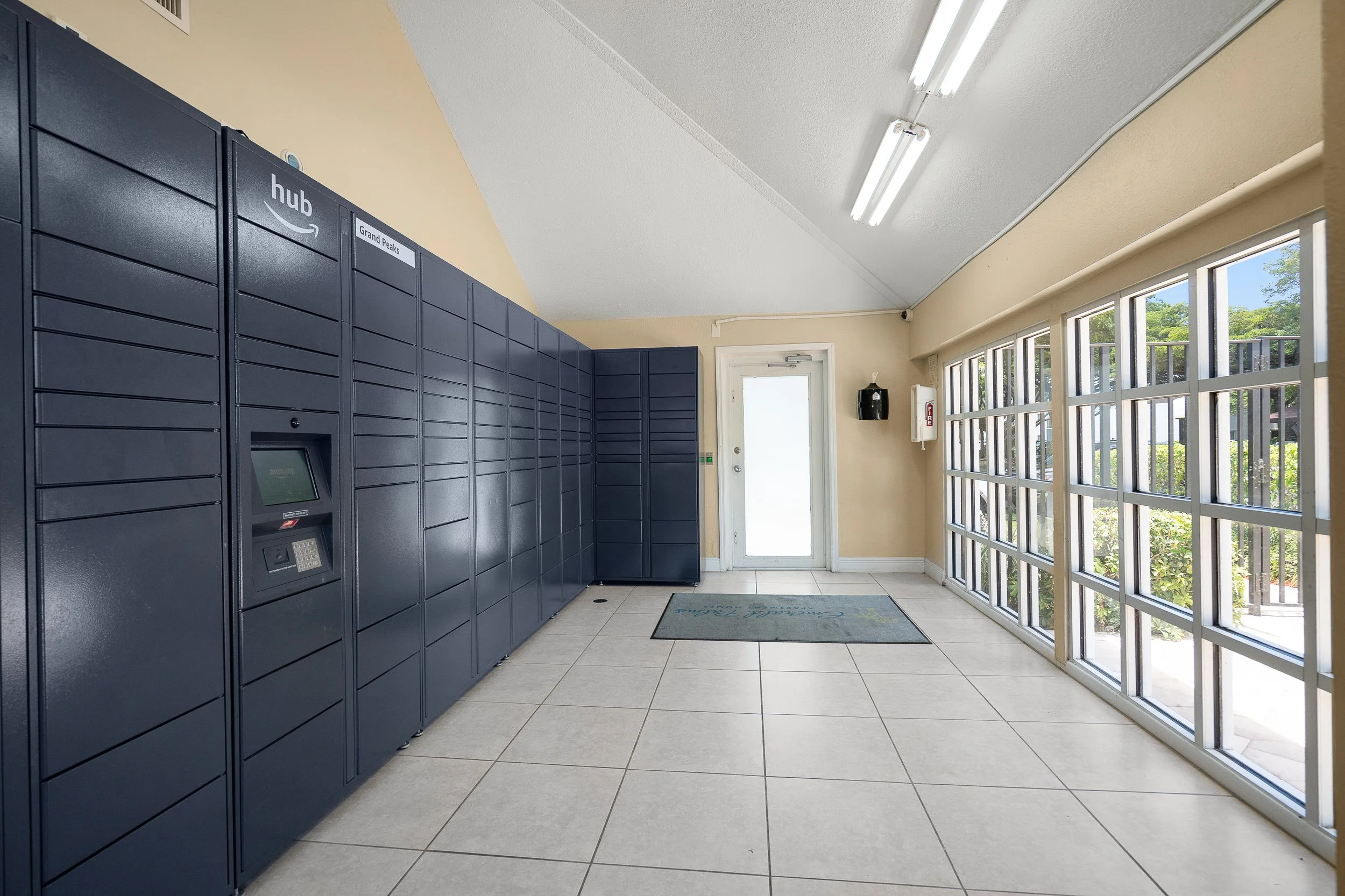 Inside a parcel locker area with a row of blue lockers on the left, a white door at the back, and large windows on the right letting in natural light.