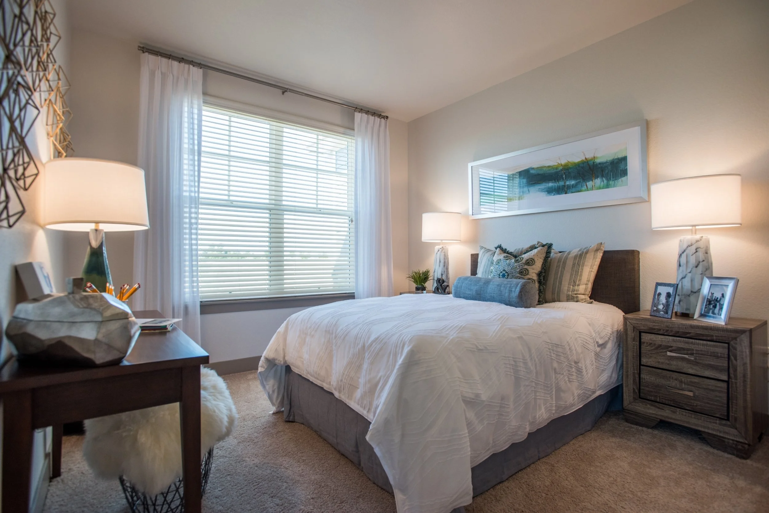 A cozy bedroom with a large bed featuring decorative pillows, two nightstands with lamps, a window with blinds and curtains, framed art above the bed, and a desk with a lamp and photos.