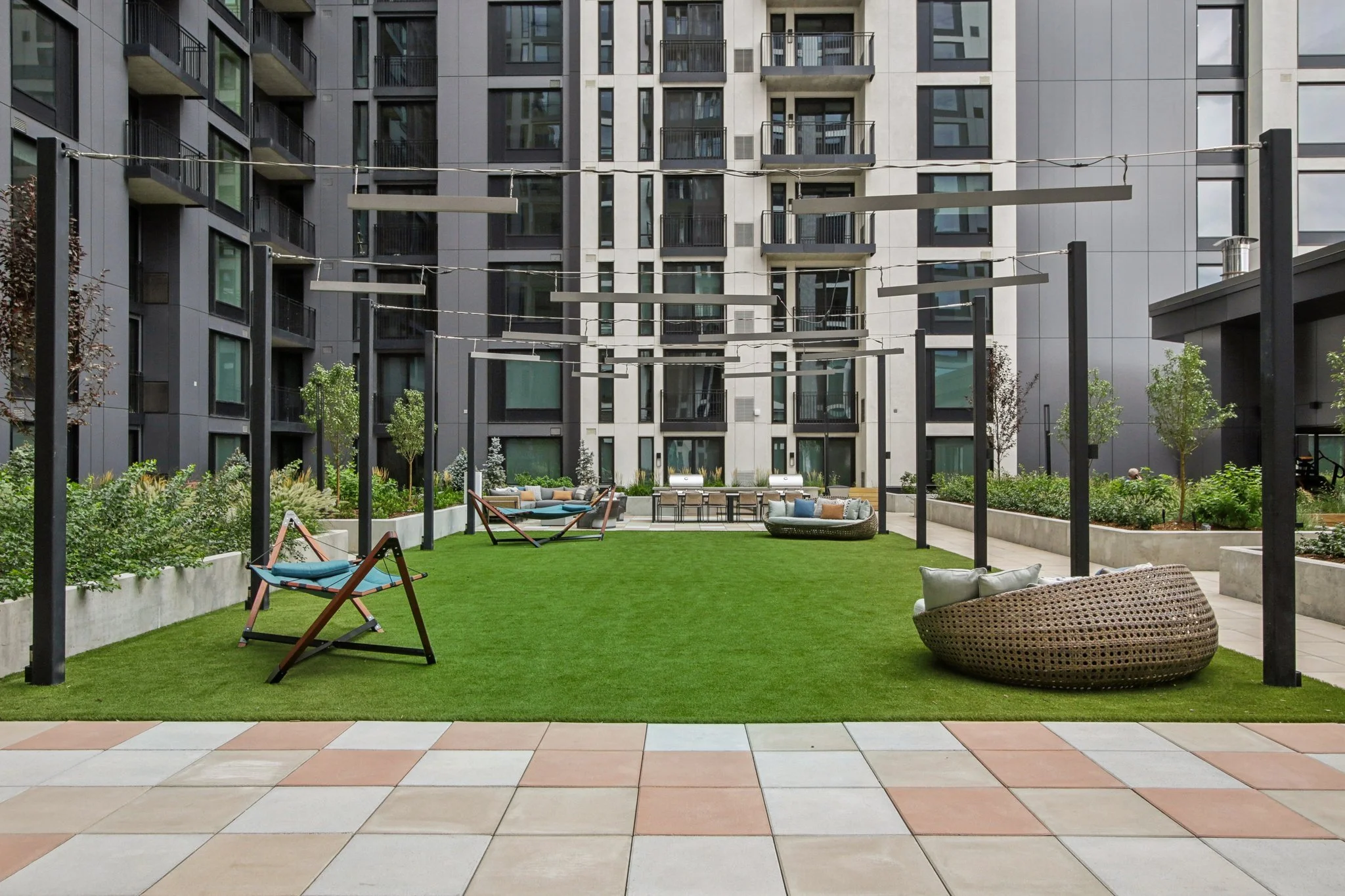 Modern rooftop courtyard with green artificial grass, surrounded by apartment buildings, featuring outdoor seating, hammocks, and decorative lighting.