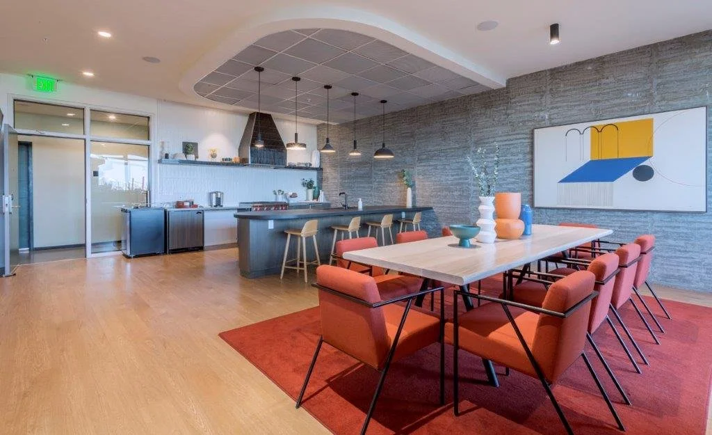 Modern lounge and dining area with a kitchen in the background. There is a large rectangular dining table with eight orange chairs. A gray countertop with four stools is also visible. The kitchen features a black range hood above a stove. The space h