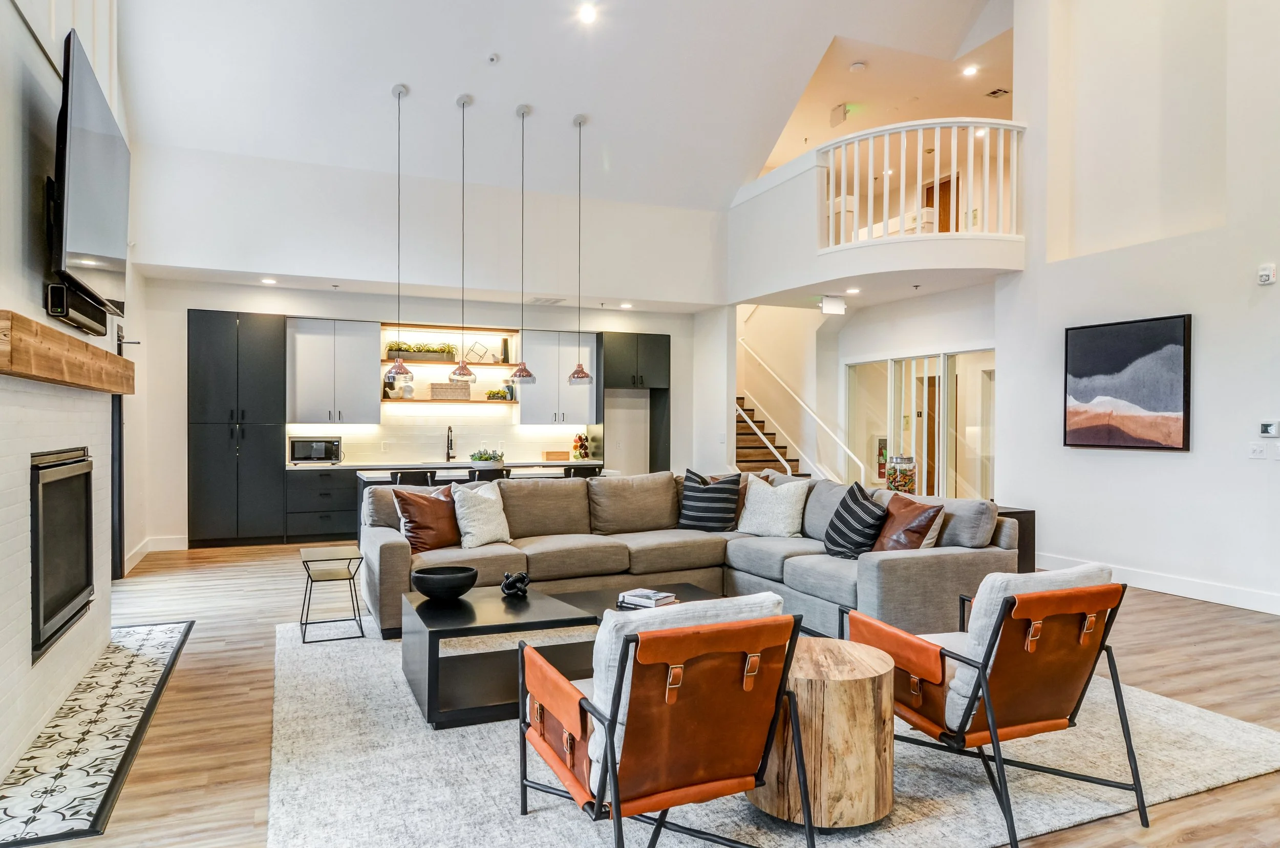 Modern living room with beige sectional sofa, black coffee table, two orange armchairs, and a fireplace. Open kitchen with black and white cabinets and pendant lights. Staircase and upper balcony overlooking the room.