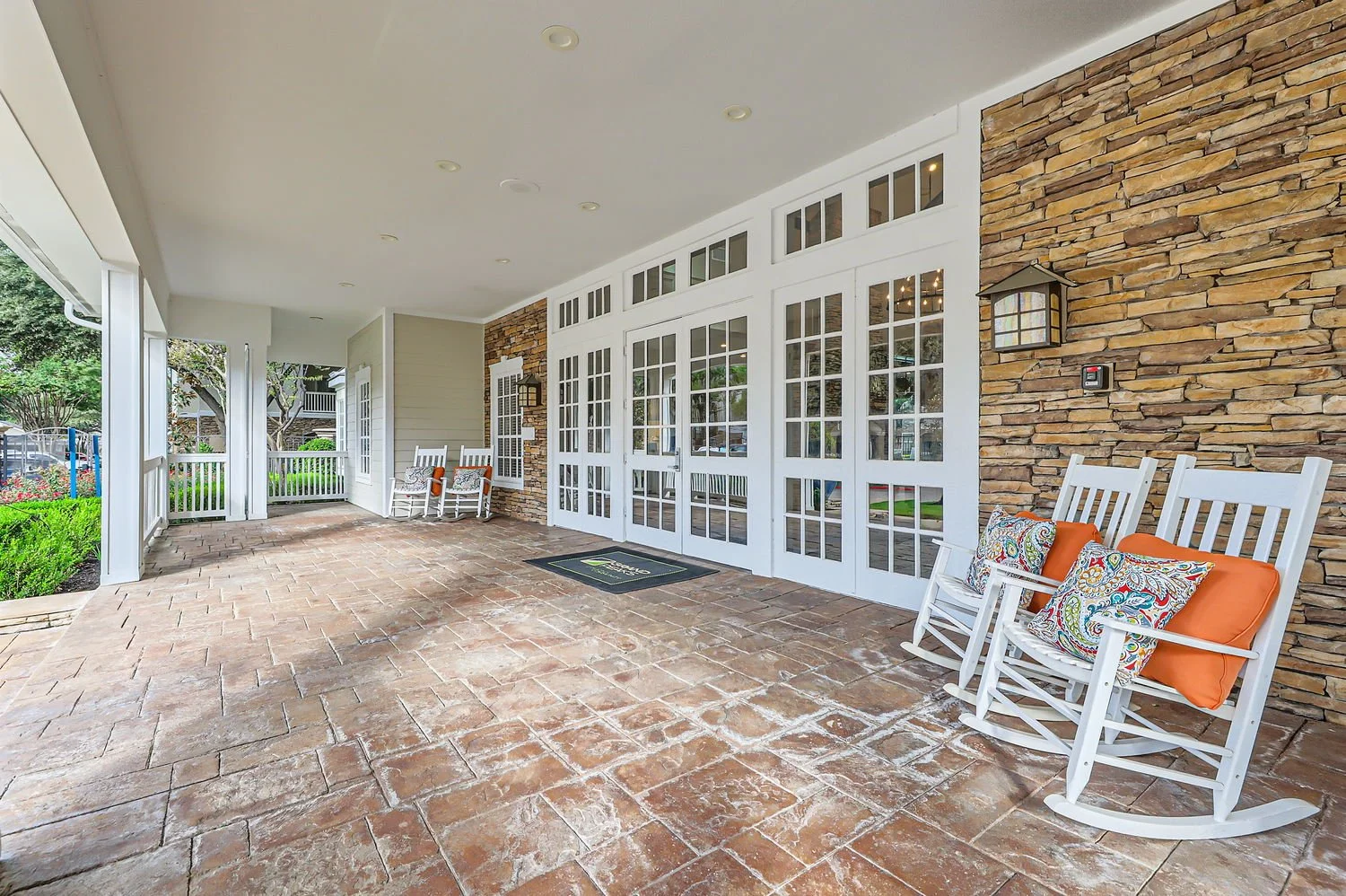 Covered porch with white rocking chairs, orange and patterned throw pillows, brick floor, and a stone wall with lantern-style lights and a door with glass panes.