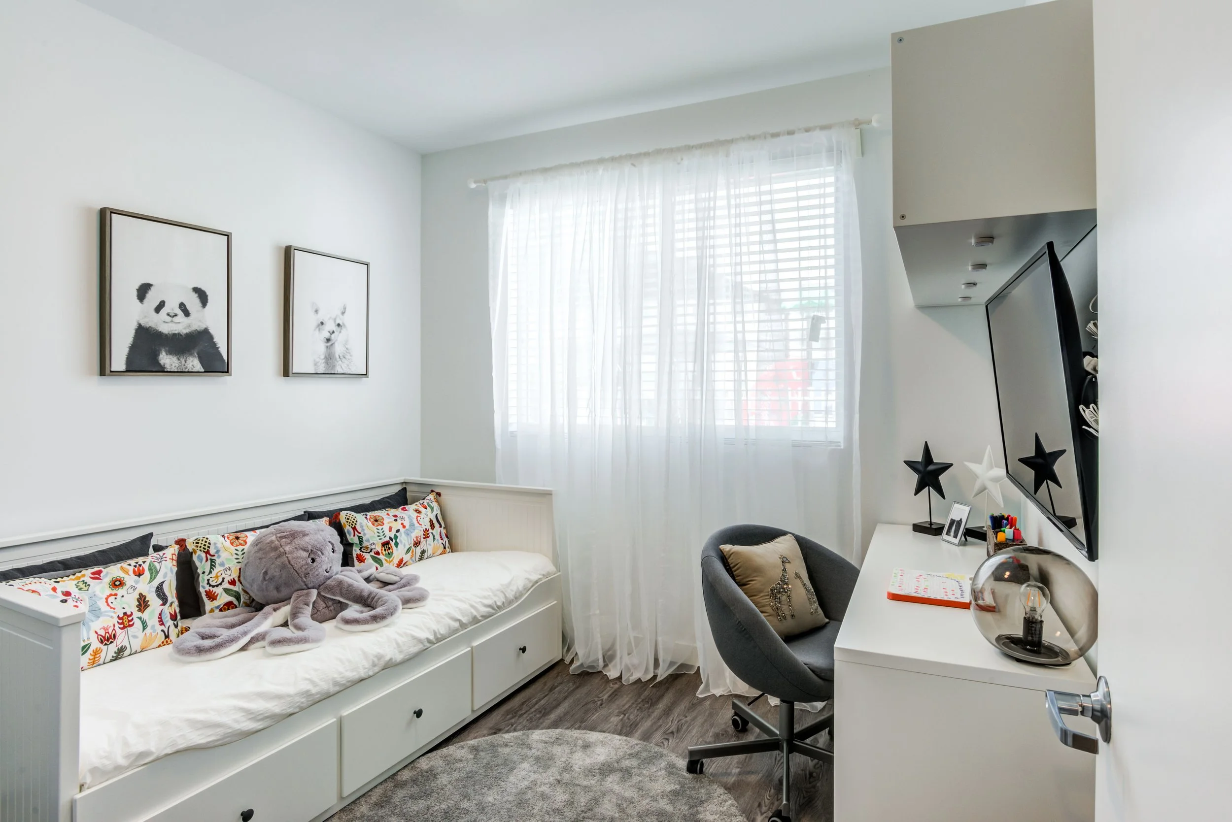 A bedroom with white walls and a window with sheer curtains. A daybed with drawer storage underneath is decorated with colorful pillows and a large plush octopus. Two framed black-and-white animal pictures hang above it. To the right, a white desk wi