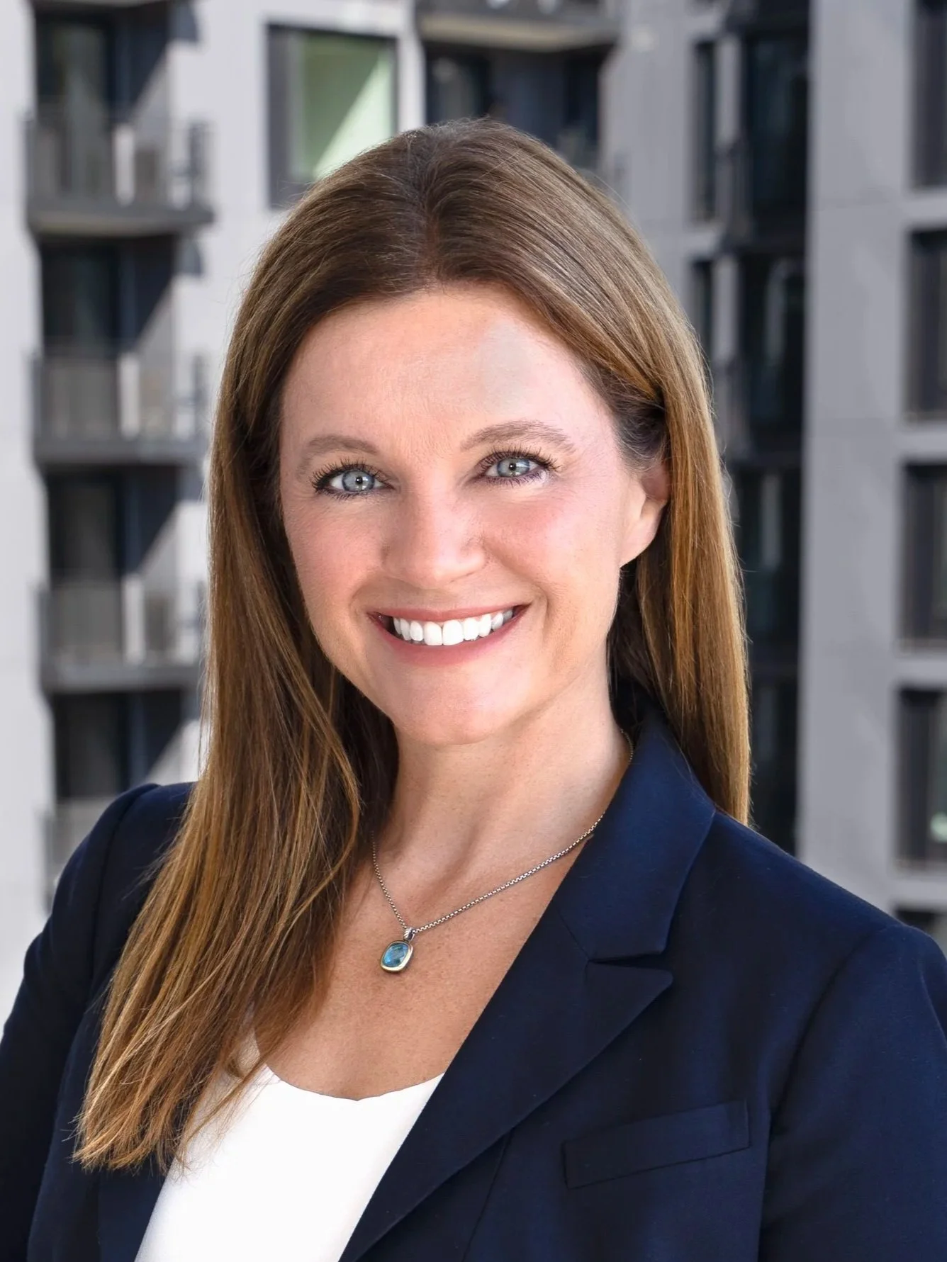 A smiling woman with long red hair, wearing a navy blazer and white top, standing outdoors in front of a modern building with balconies.