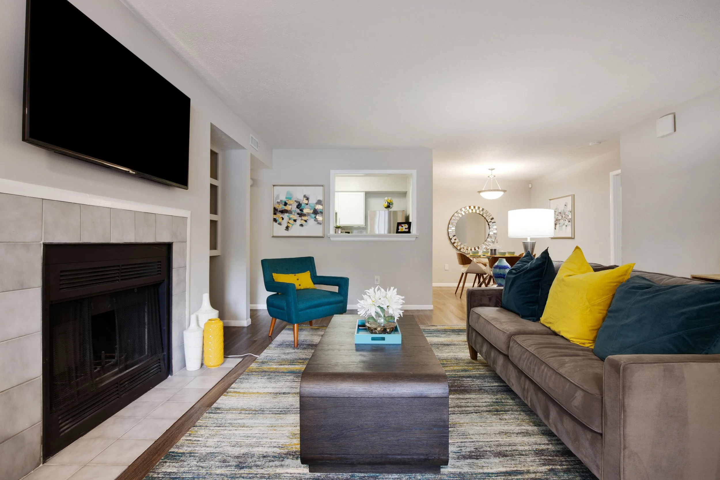 Living room with a fireplace, a wall-mounted TV, a brown sofa with yellow and blue pillows, a wooden coffee table with a floral decoration, a blue armchair with a yellow pillow, and a dining area with a round mirror and artwork on the walls.