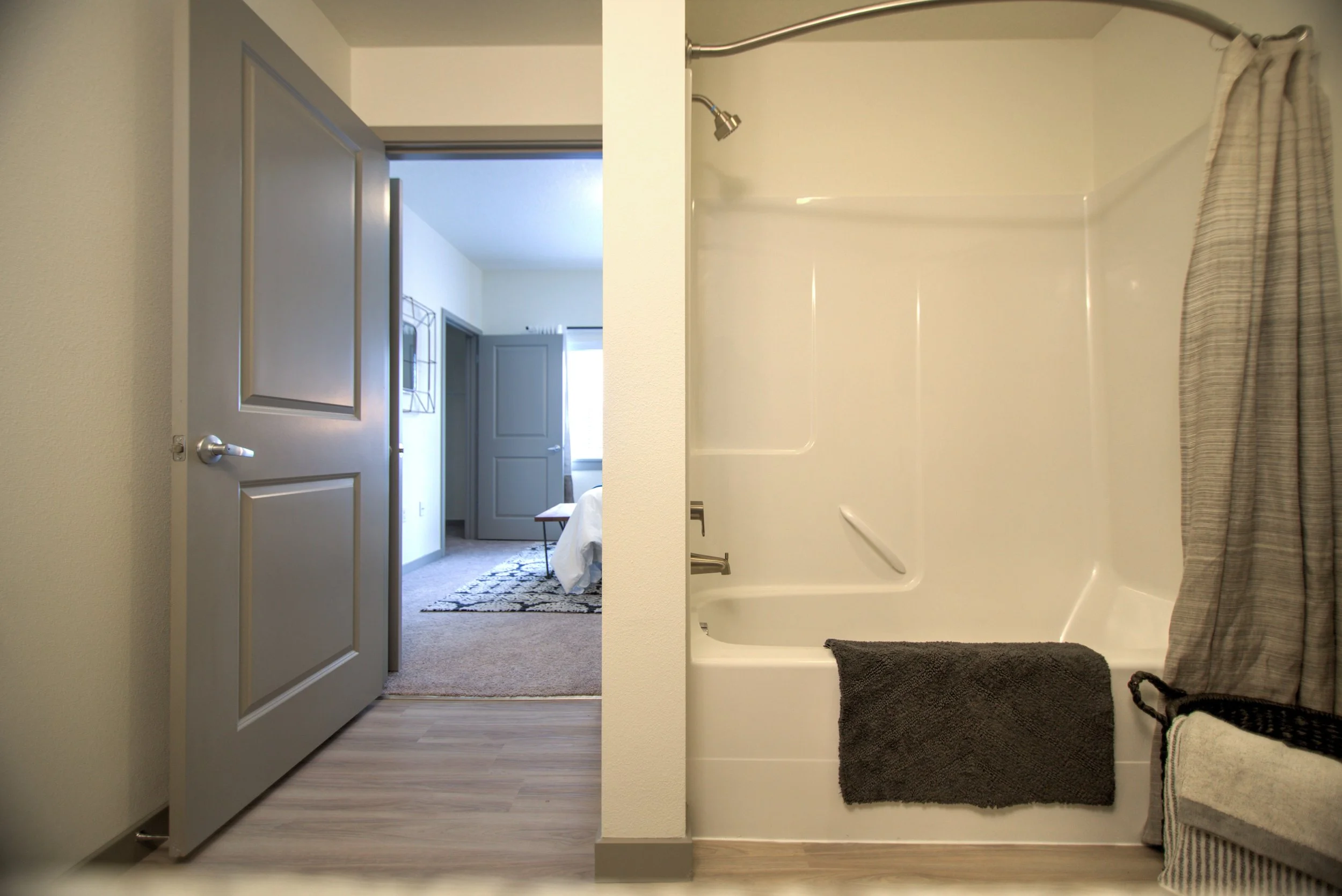 The image shows a bathroom with a bathtub and shower on the right, with a dark towel hanging over the side. To the left, there is an open door leading to a bedroom with gray walls, a window, and a bed. The bathroom has light-colored walls and wood la