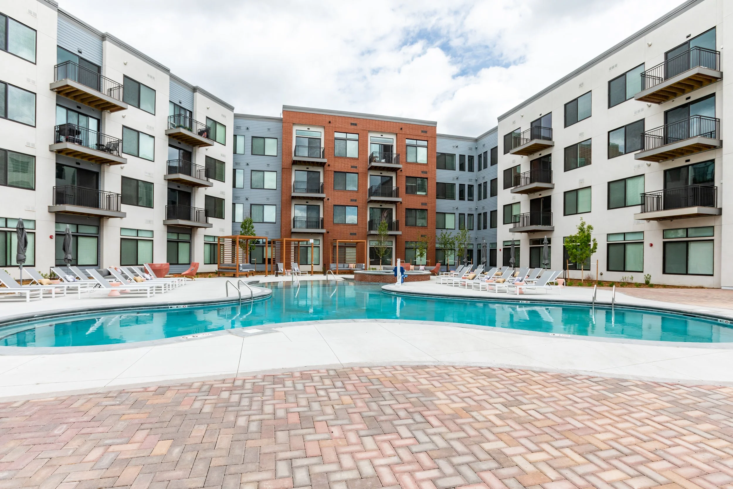 Modern apartment complex with a central swimming pool, surrounded by lounge chairs and small trees.