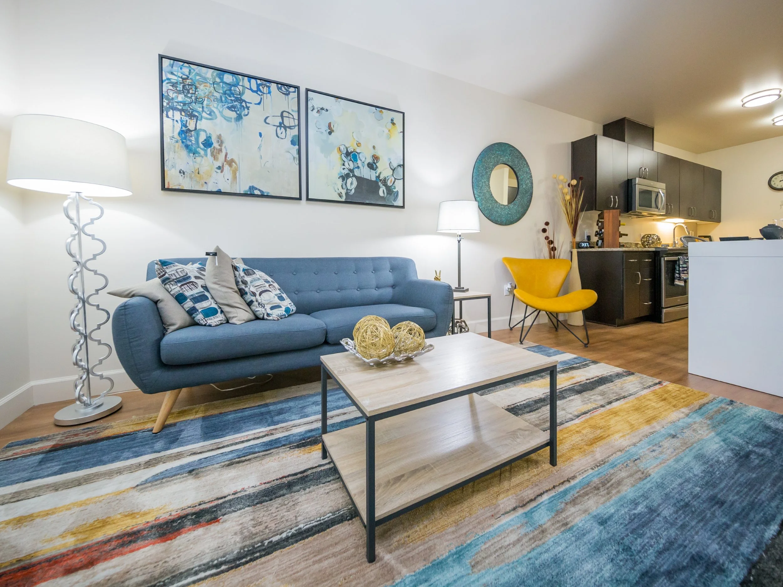 Living room with blue sofa, yellow chair, modern artwork, and kitchen in background.