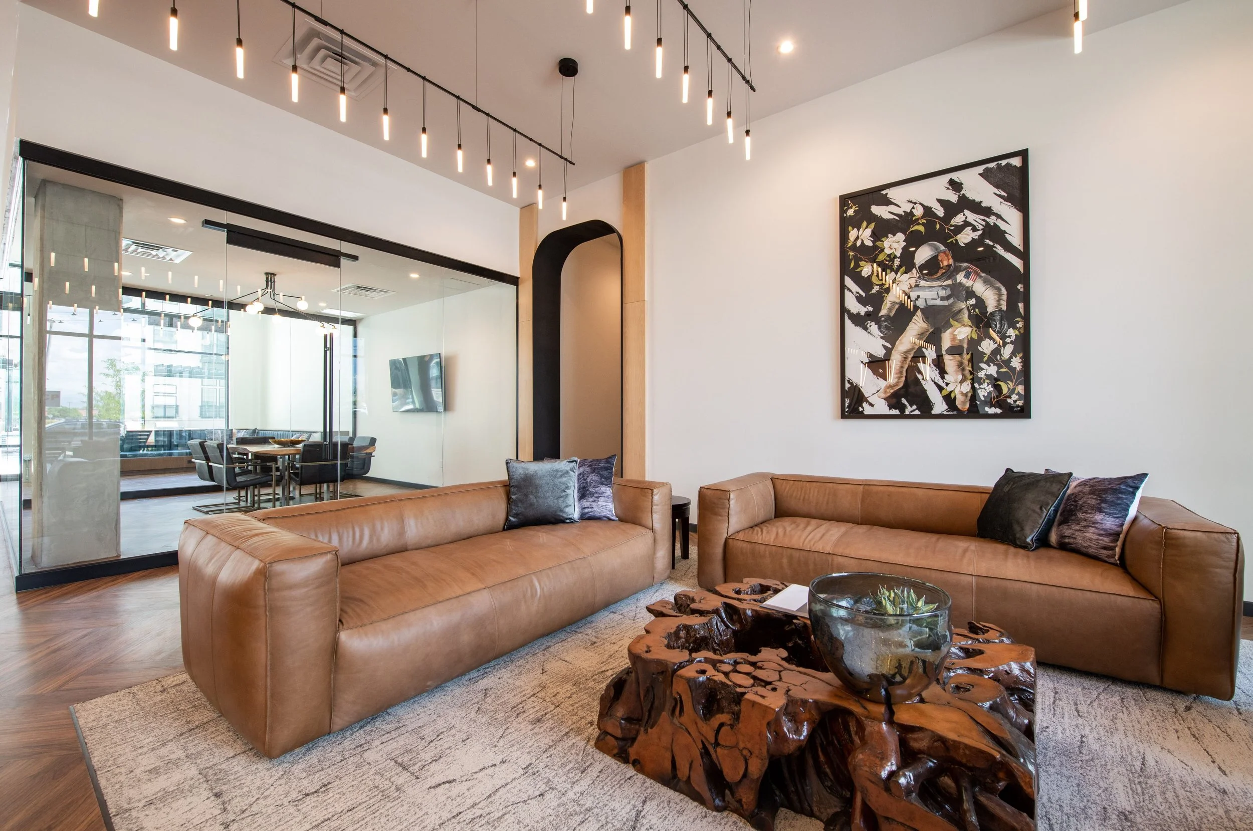 Modern office lounge with two brown leather sofas, decorative pillows, a rustic wooden coffee table, abstract astronaut artwork on the wall, and a glass wall separating the lounge from a conference room with a large window and a dining table.