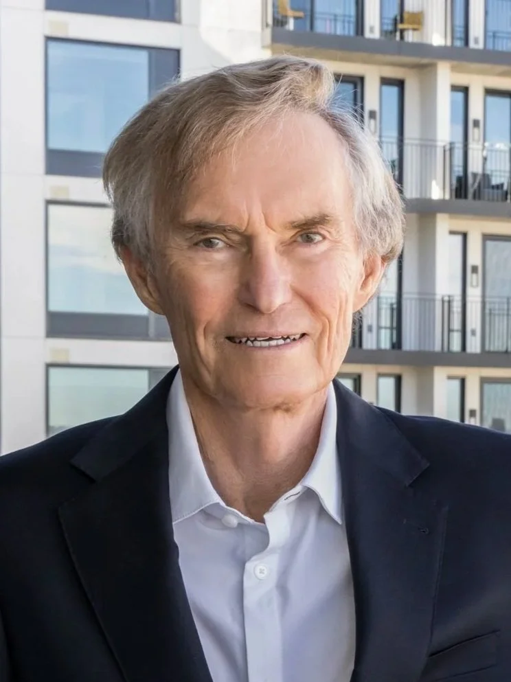 A mature man with light-colored hair wearing a white shirt and black jacket, standing outdoors in front of a modern apartment building.