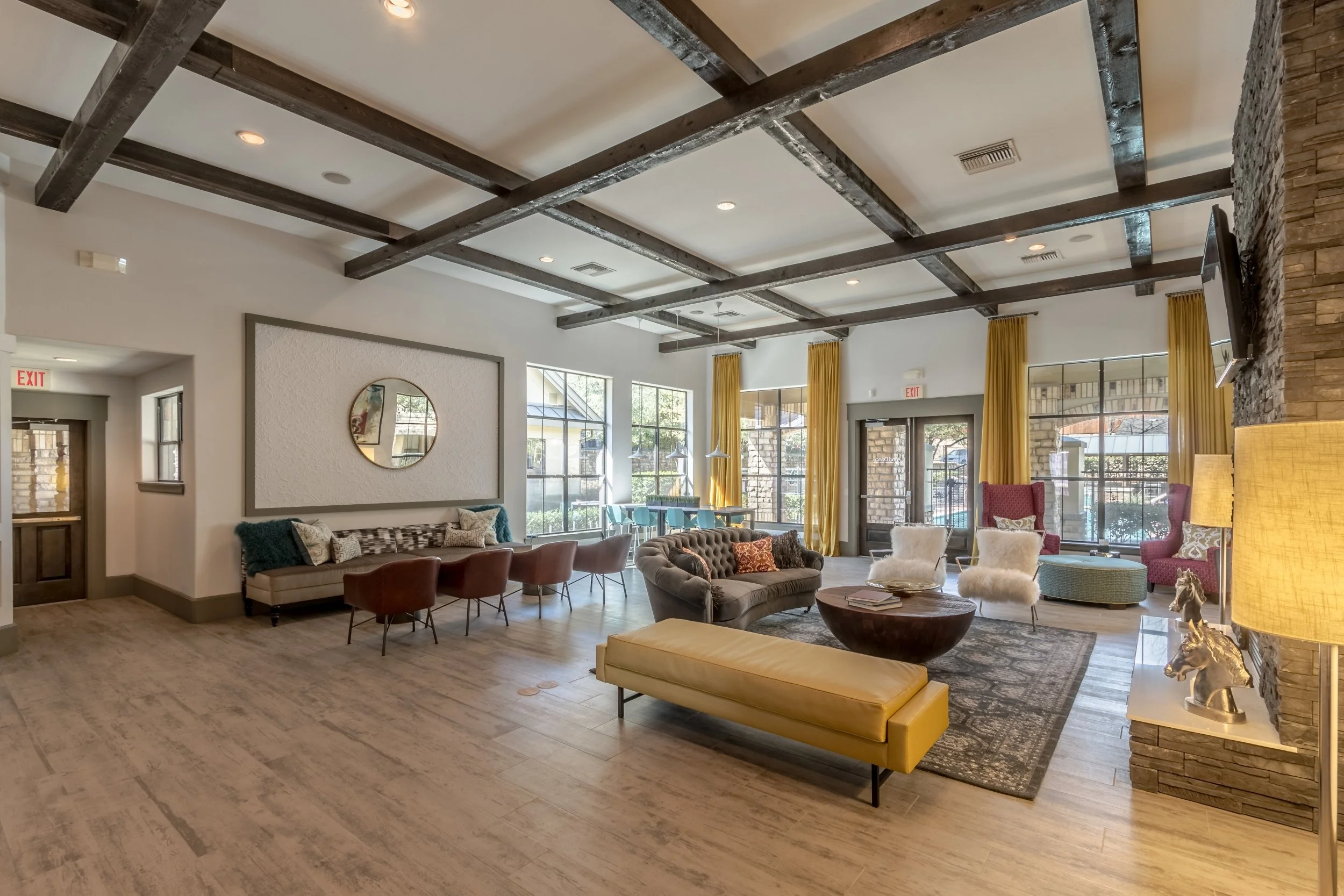 A spacious and modern lounge area with various seating options, large windows with yellow curtains, a stone accent wall, and wooden ceiling beams.