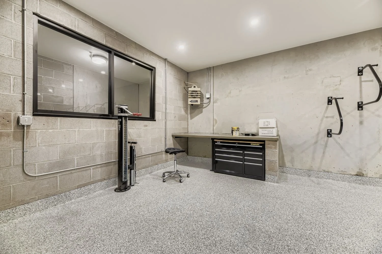 Empty garage space with concrete block and wall surfaces, a workbench with tools and storage drawers, a stool, and wall-mounted shelves.
