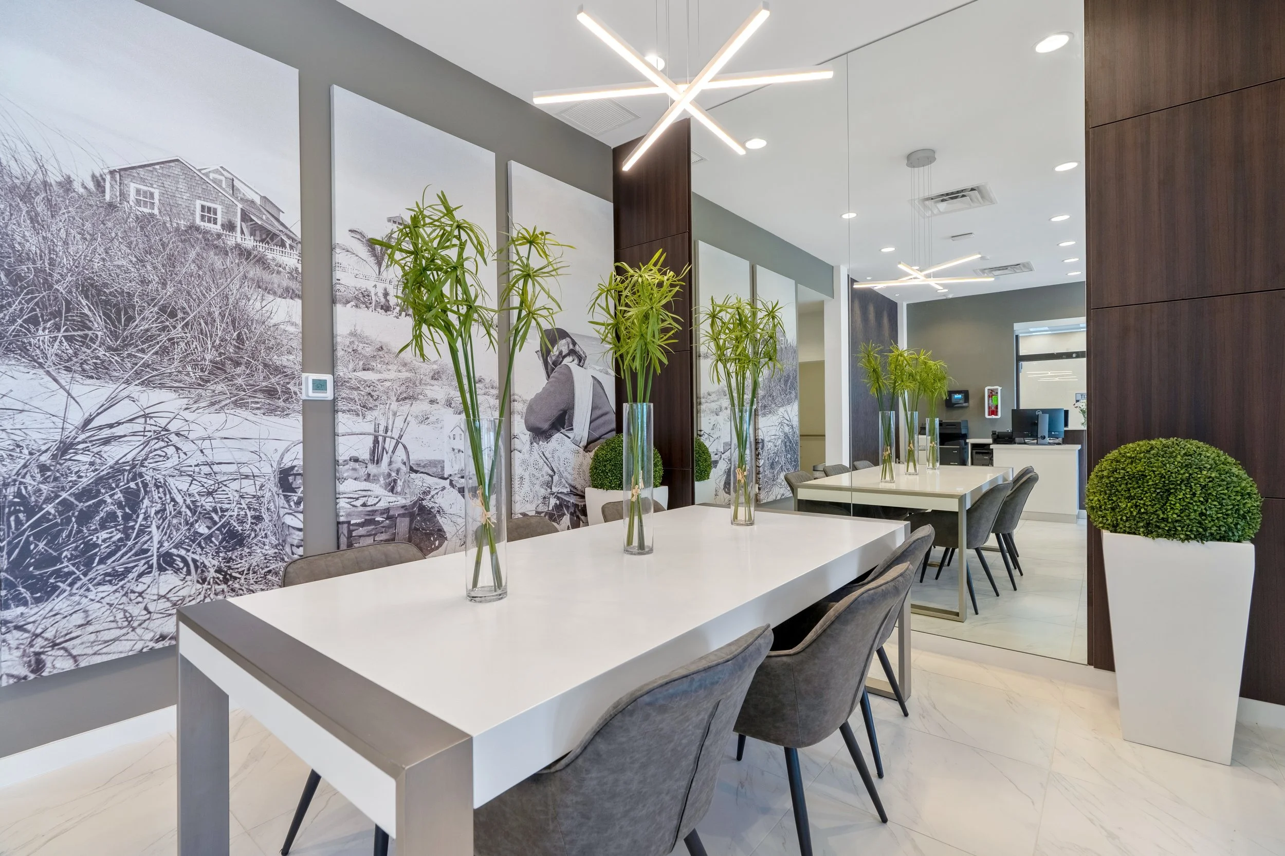 Modern office or meeting room with white tables, gray chairs, tall green plants in glass vases, large black-and-white landscape photographs on the wall, contemporary ceiling lights, and a reception area with a computer and phone.