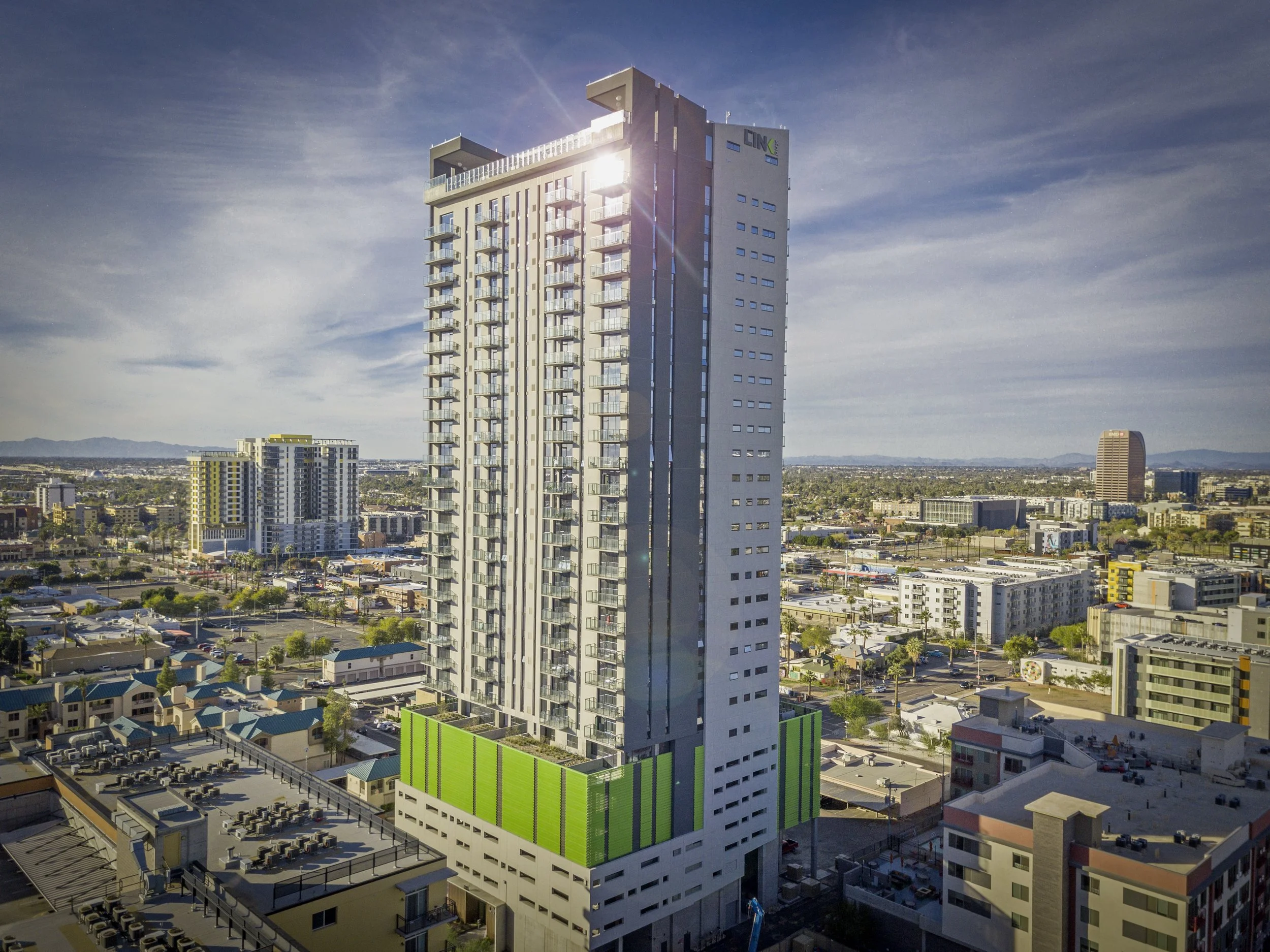 A tall modern high-rise building with a green and white facade at the base, located in an urban area with other buildings and parking lots visible, under a partly cloudy sky with sunlight reflecting off the building.