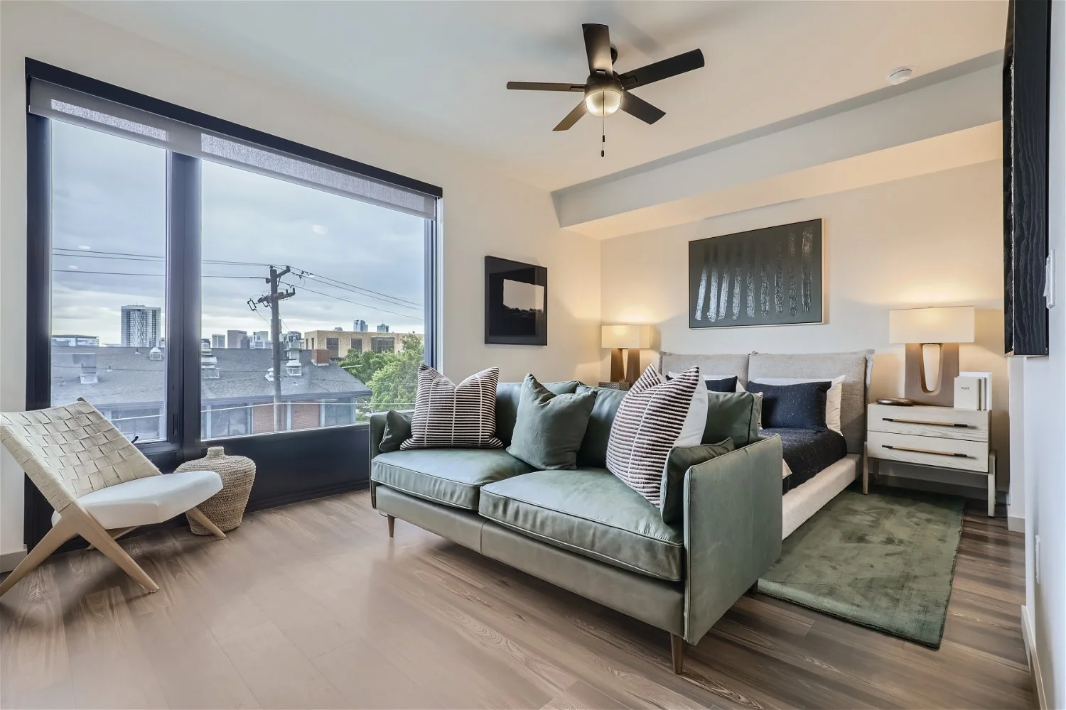 Modern living room with large window, green sofa, corner chair, bed with black and white bedding, and nightstand with lamps