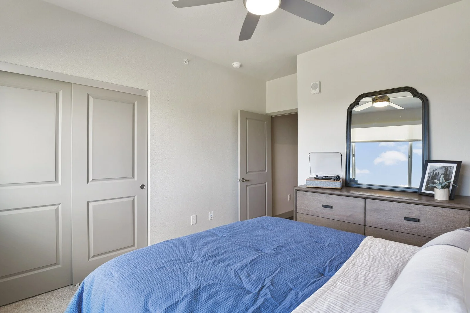 A bedroom with a bed covered in a blue quilt, a dresser with a mirror, a record player, a framed picture, a plant, and a window showing blue sky with clouds.
