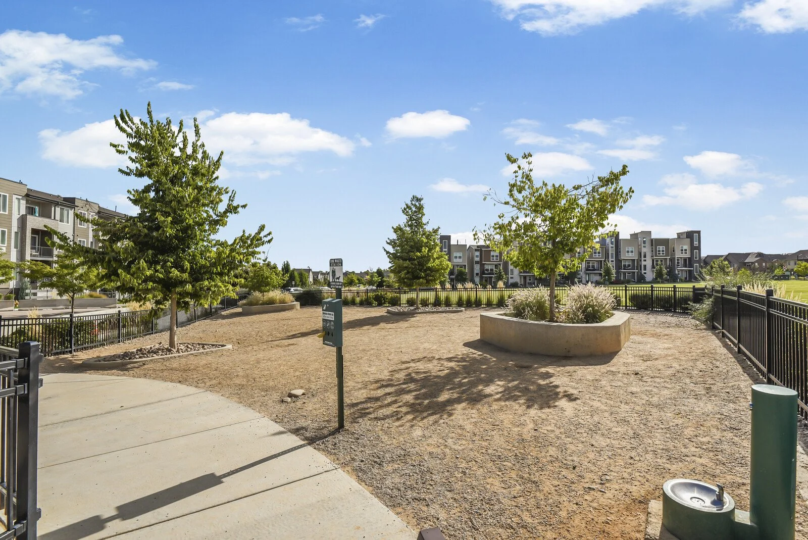 A small public park with three trees in raised concrete planters, surrounded by metal fencing, with apartment buildings in the background, under a sunny sky with a few clouds.