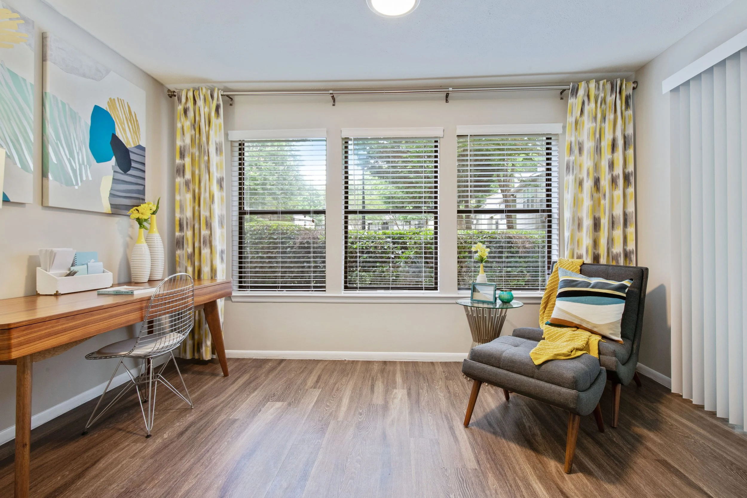 Bright living room with three large windows featuring horizontal blinds, decorated with yellow and gray patterned curtains. Contains a wooden desk with two white vases and flowers, a transparent wire chair, a gray lounge chair with pillows and a yell
