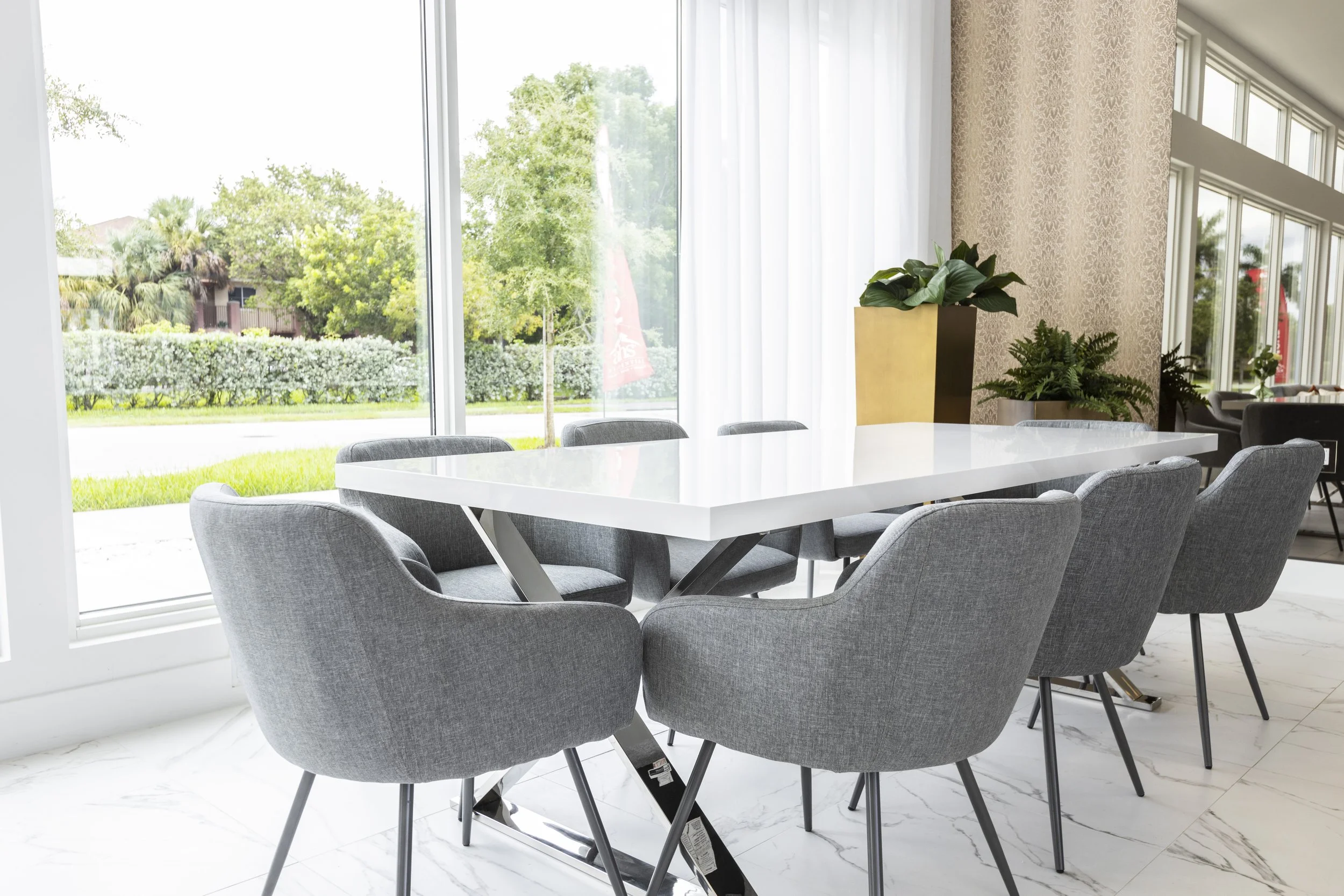 Modern dining area with a white table surrounded by eight gray upholstered chairs, large windows with white curtains, and green plants on a marble floor.