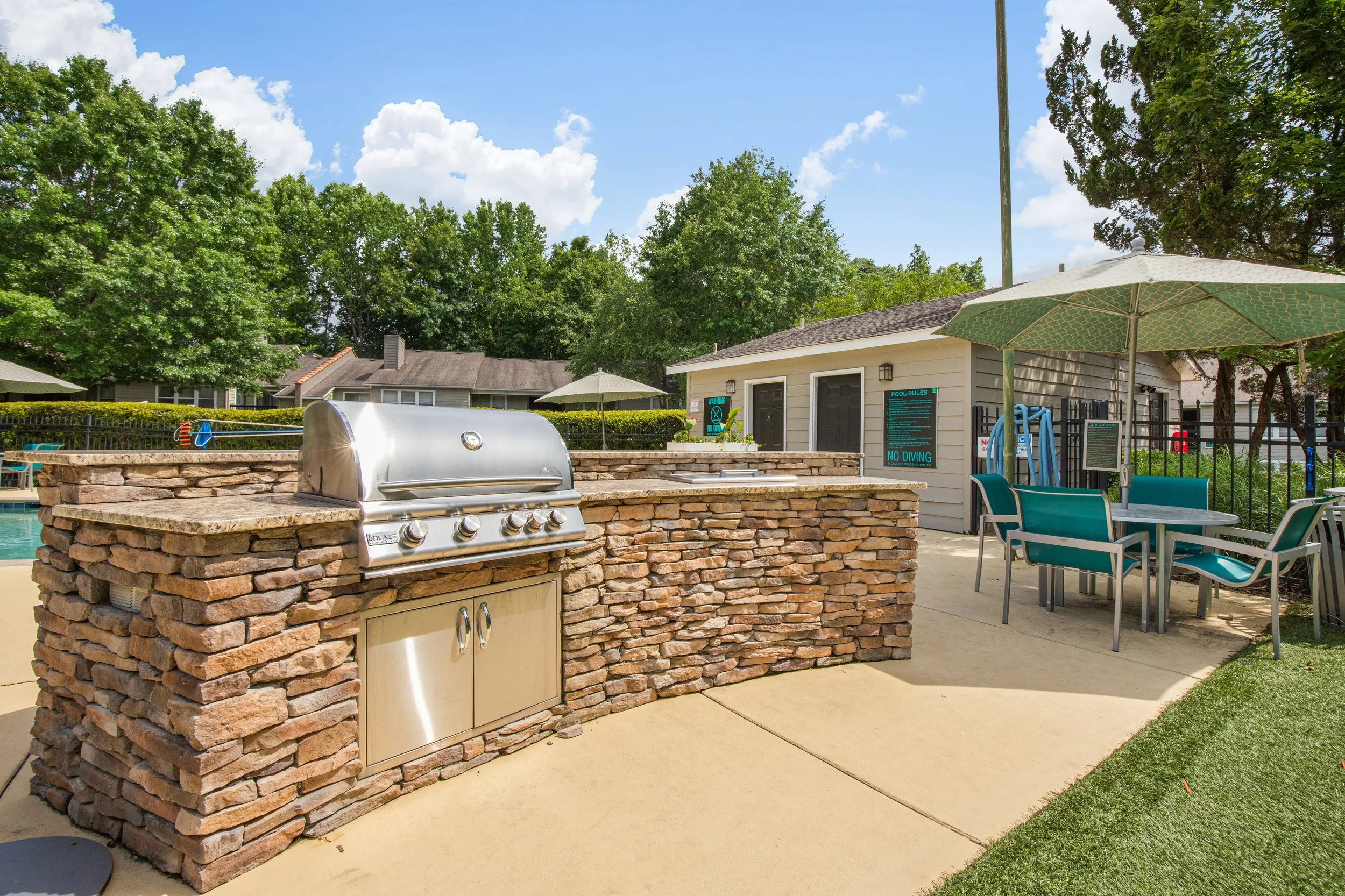 Outdoor swimming pool with a built-in grill station, patio furniture, umbrellas, and a pool house surrounded by trees and a clear blue sky.