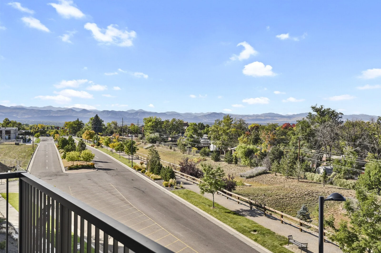 A view from a balcony showing a paved parking lot with no cars, surrounded by trees, grass, and a sidewalk. In the background, there are distant mountains and a partly cloudy blue sky.