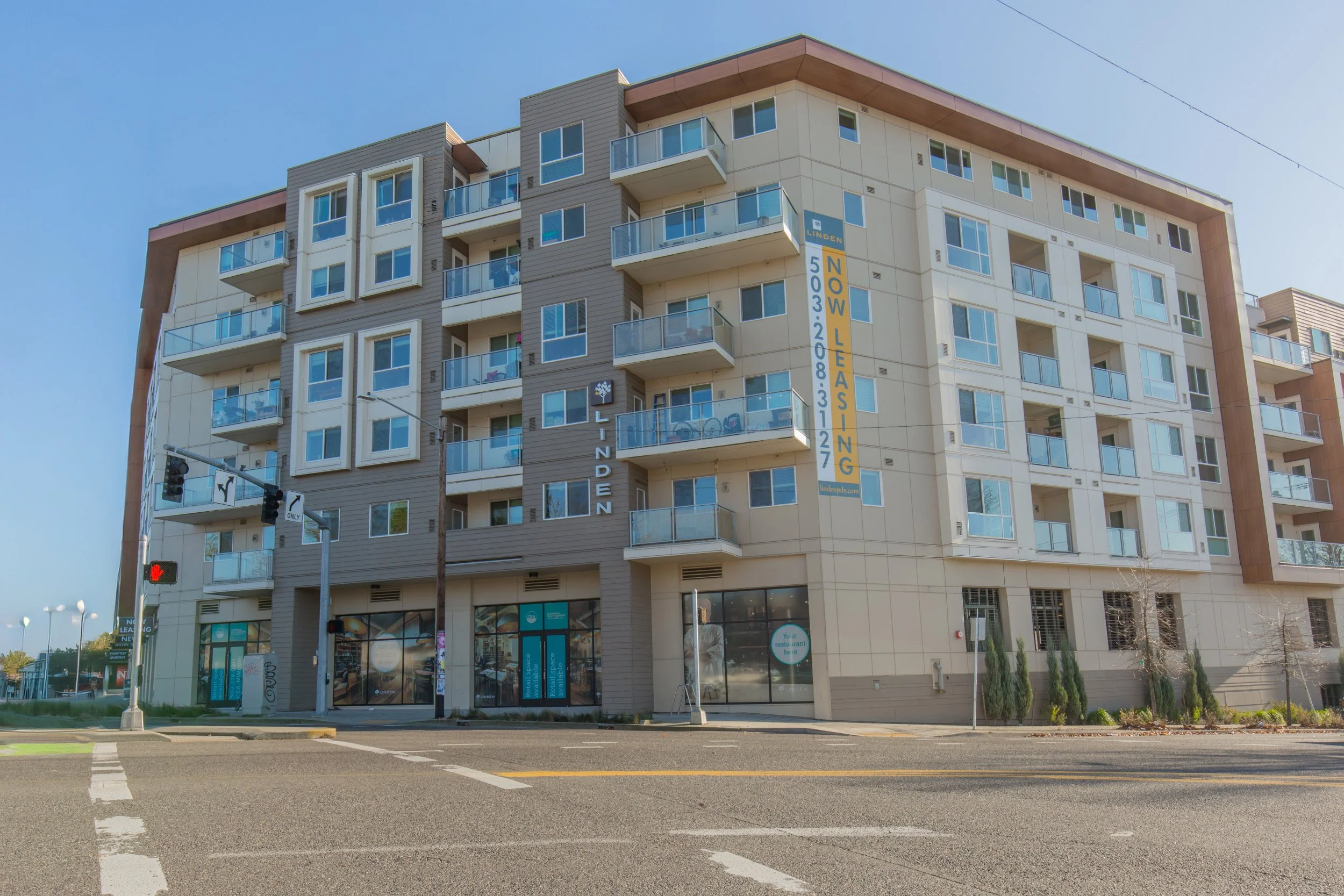 Modern multi-story apartment building with balconies, large windows, and a banner advertising leasing, on a city street with traffic lights and crosswalks.