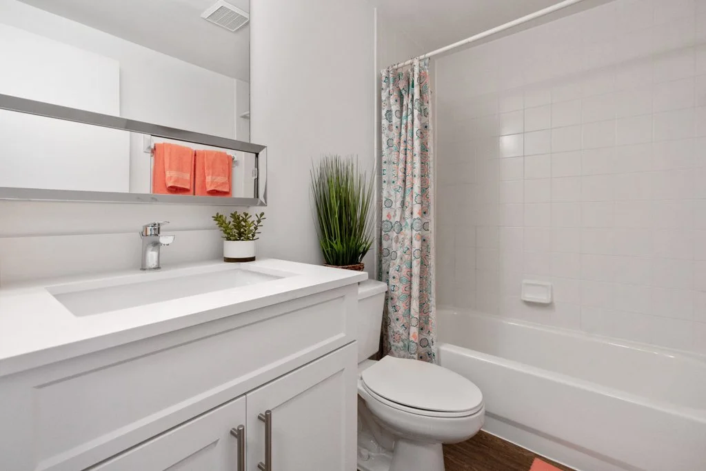 Small modern bathroom with white vanity, gray mirror, coral towels, potted green plant, bathtub with patterned shower curtain, and wood-textured floor.