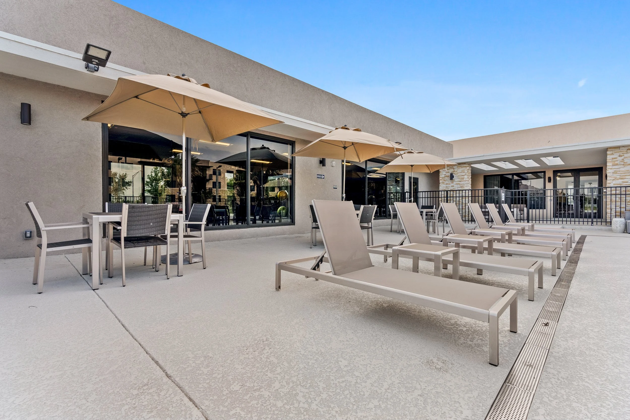 Outdoor patio area with lounge chairs, tables, and umbrellas in front of a modern building with large glass windows.