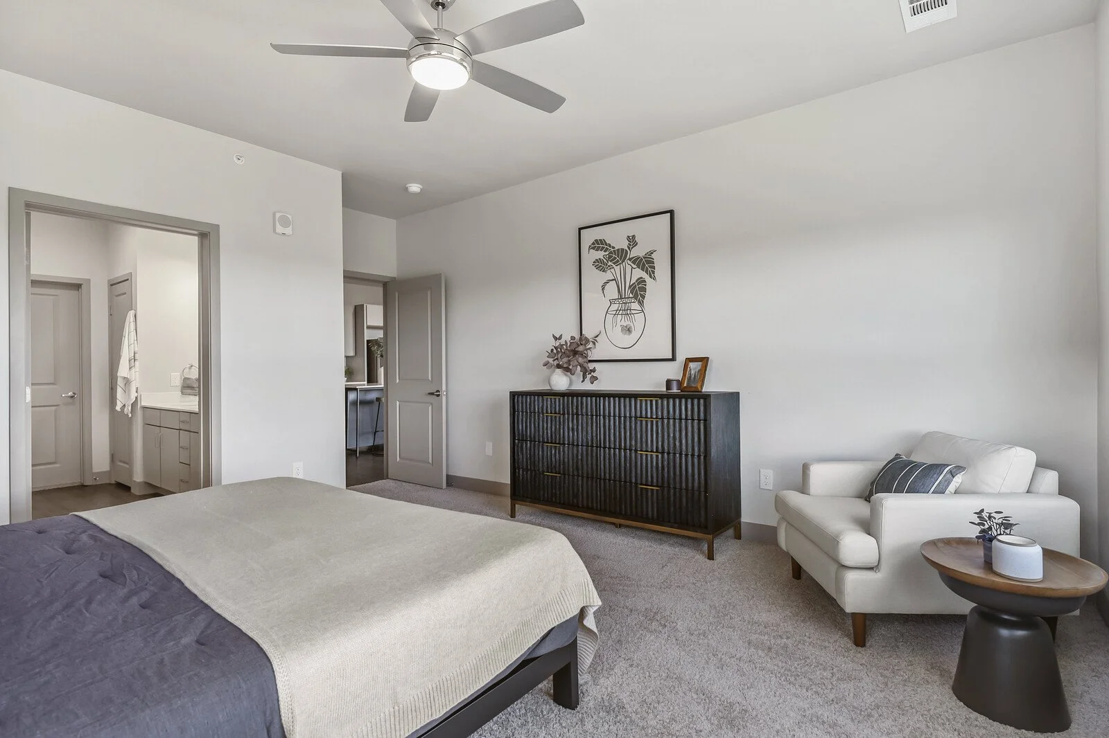 A modern bedroom with a large bed, white walls, a ceiling fan, a black dresser with decorative items, a white armchair, and a small wooden side table.