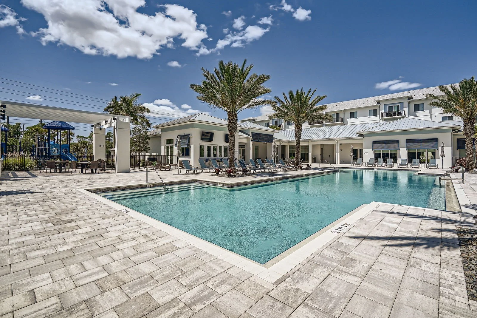 A swimming pool area at a resort or complex with lounge chairs, palm trees, a playground, and a modern building in the background under a partly cloudy sky.